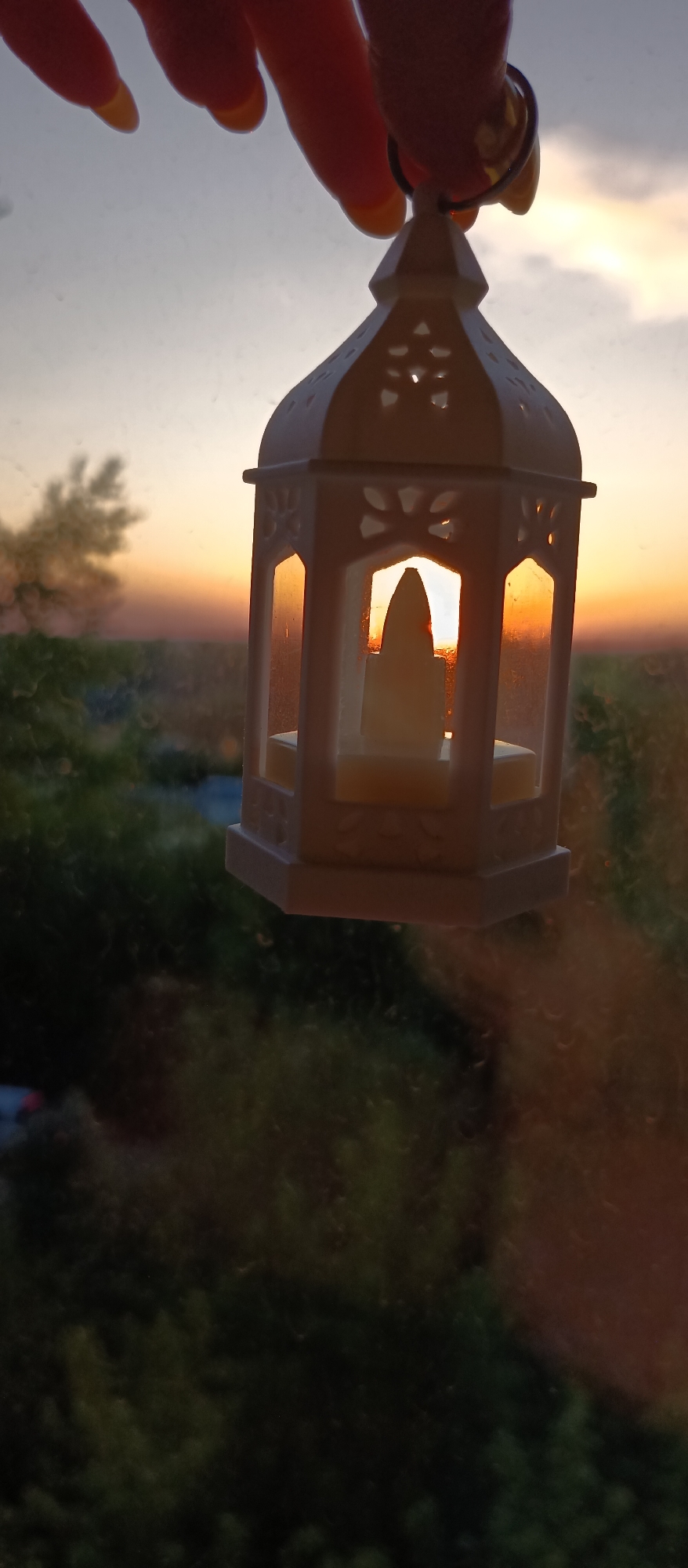A hand with yellow-painted nails holds a small, white, decorative lantern against the backdrop of a sunset. The lantern contains a small, white, flameless candle. The blurred background shows greenery and buildings under a sky with an orange glow from the setting sun. The view appears to be seen through a window with droplets.