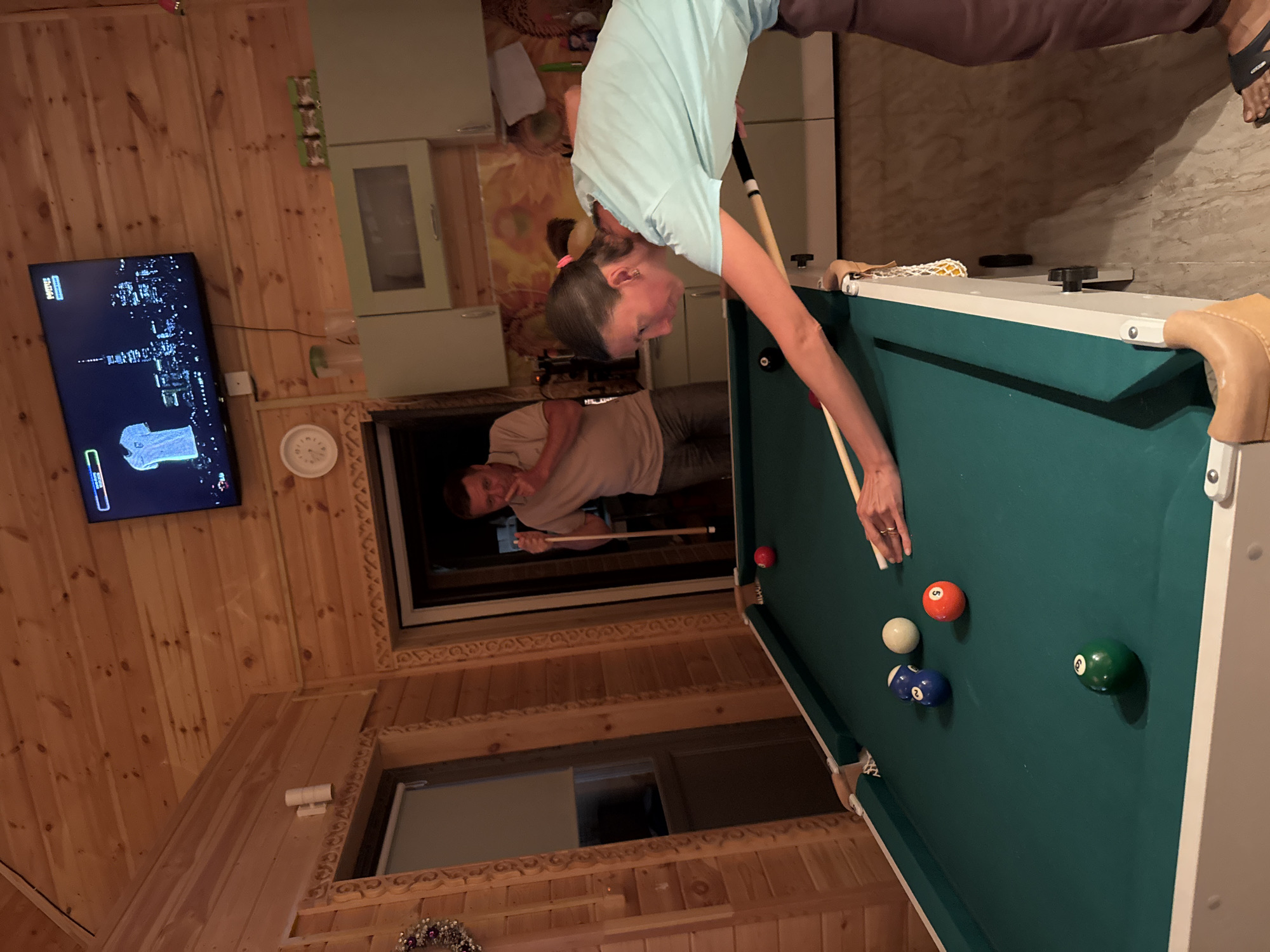 In Zelenaya Roshcha, Russia, a woman in a light blue t-shirt is playing pool indoors in a room with wooden walls. She is leaning over the green pool table, cue in hand, focused on a shot. Nearby, a man in a light brown t-shirt stands with a pool cue, appearing to be watching or waiting his turn. The room features a television mounted on the wall displaying an image of a city skyline and a white shirt, along with kitchen cabinets and a clock on the wall. Several pool balls are scattered across the table, including a red ball, a white ball, and two blue balls marked with numbers.