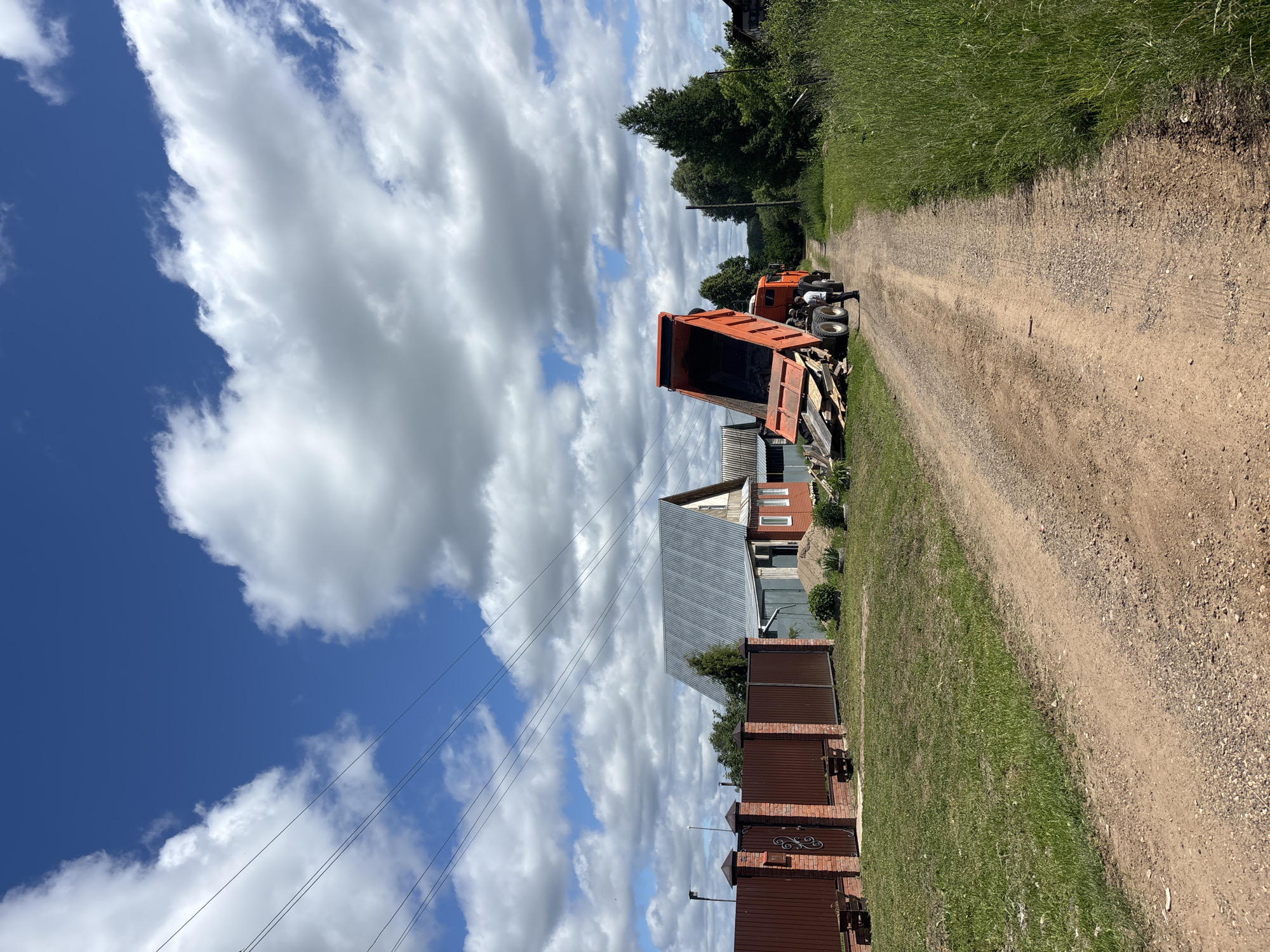 An orange dump truck is parked on a dirt road in a rural area, likely in the city of Niva, Russia, with its bed raised and partially emptied of materials. A person in a white shirt and dark pants stands near the rear of the truck, facing away from the camera. The truck is positioned next to a pile of wood and debris. To the left of the road, there is a fenced property with a brick fence and gate, leading to a house with a metal roof. To the right of the road, tall green grass and trees line the path. The sky is blue with scattered white clouds. The scene suggests construction or cleanup activity is taking place.