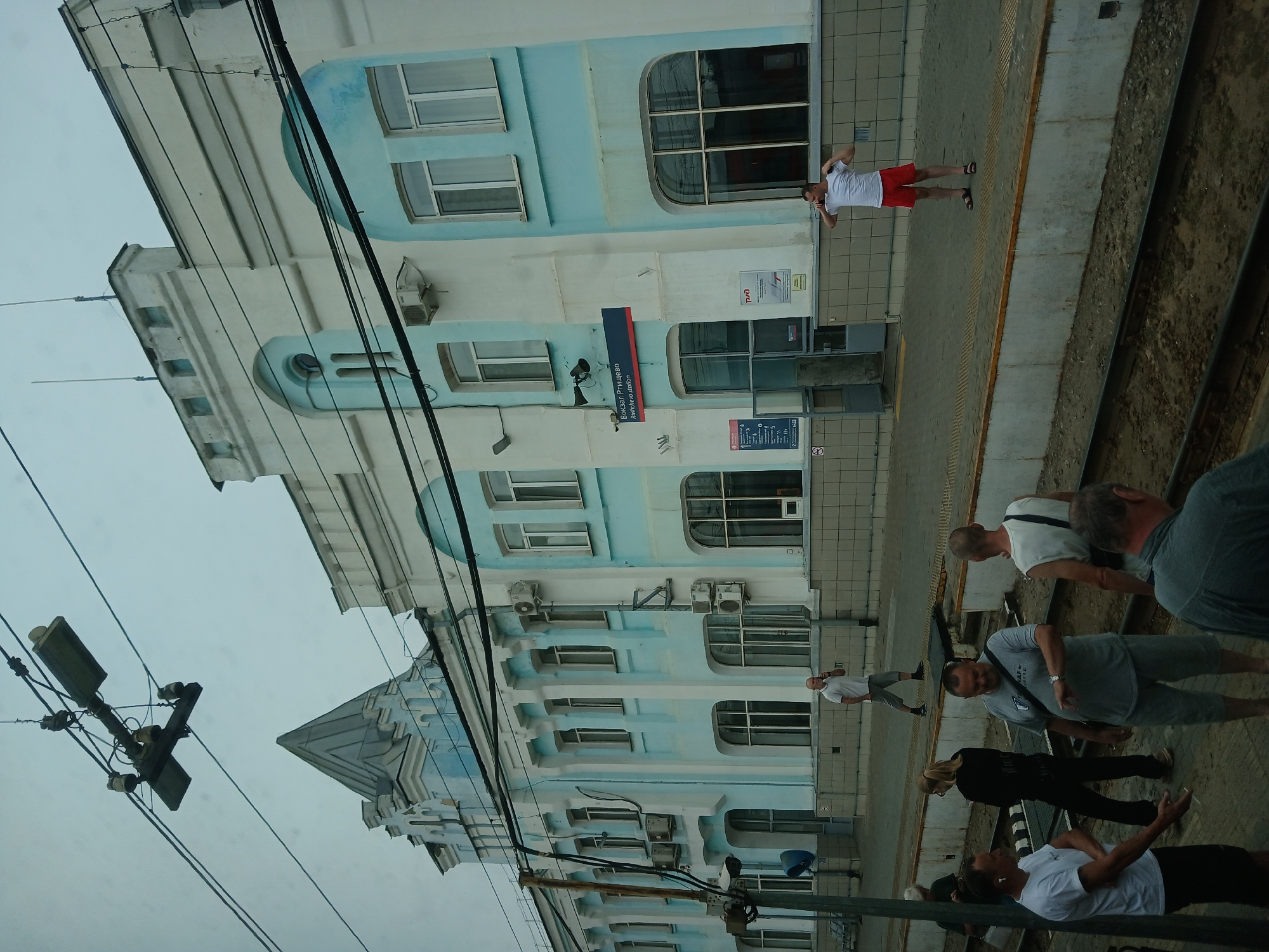 The image depicts the Rtishchevo railway station in Rtishchevo, Russia, under an overcast sky. The station building, painted in a light blue and white color scheme, features multiple windows, some with arched tops, and decorative architectural elements. Overhead, a complex network of electrical wires and a streetlamp structure are visible, common for railway infrastructure.
On the platform, several individuals are present. A man in a white t-shirt and red shorts stands near the building, holding a phone to his ear. Further down the platform, a group of people are gathered, some appearing to be waiting or conversing. One man in a gray t-shirt and shorts is looking at his watch, while another man in a white t-shirt stands with his hands in his pockets. A woman in black clothing is also part of the group.
The foreground shows railway tracks and the platform edge. The overall scene conveys a typical operational day at a Russian railway station.