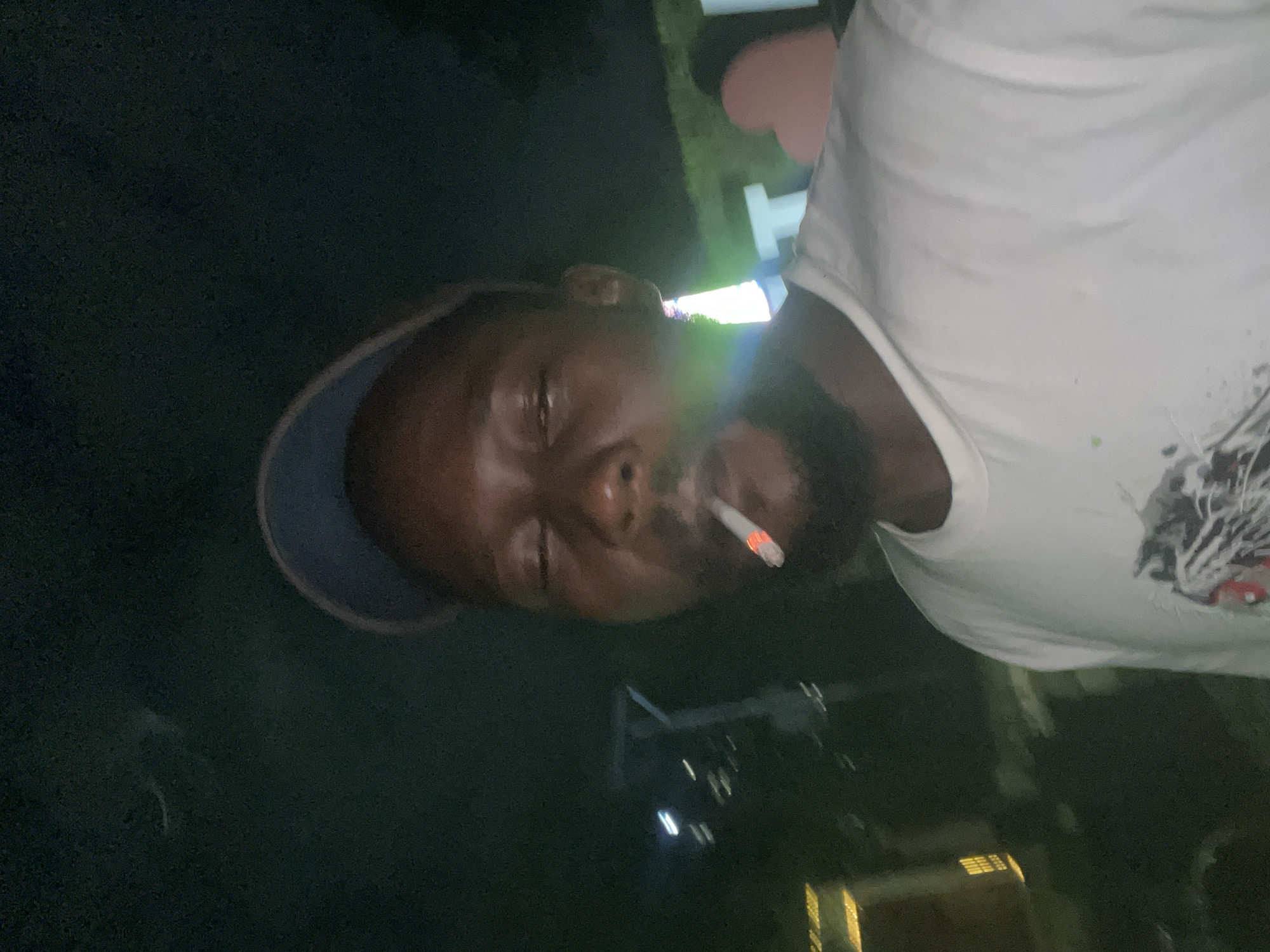 A man wearing a white t-shirt and a blue baseball cap is smoking a cigarette at night. He is looking towards the camera with his eyes closed. In the background, there are blurry lights from buildings and some illuminated signs. The scene appears to be outdoors in an urban environment. The provided crops do not offer any identifiable landmarks or specific details that would allow for the precise location within Abuja, Nigeria to be determined.