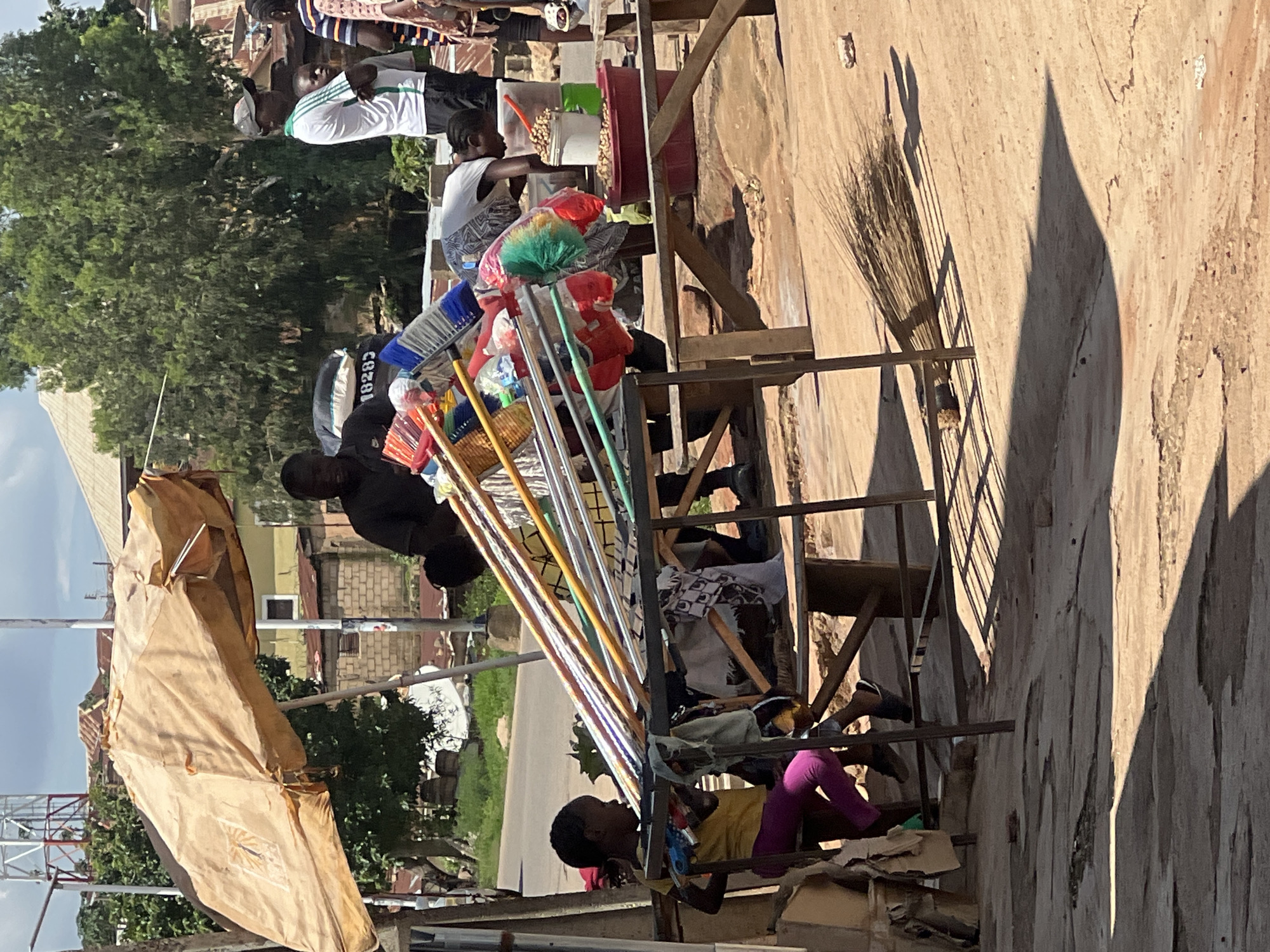 This image captures a street scene in Jos, Nigeria, likely in a market or commercial area, characterized by its dusty, unpaved ground and modest structures. A stall, partially shaded by a weathered, light brown umbrella, displays a collection of colorful brooms and brushes with plastic handles. A young girl, wearing a yellow t-shirt and purple leggings, sits on a stool at the front left of the stall, her gaze directed to the right. Behind her, a rack of brooms and brushes is prominently featured. Several adults are also present. One man in a dark shirt stands behind the stall, while another person, wearing a white t-shirt with green trim and a baseball cap, stands on the right side of the frame, arms crossed. A woman in a patterned dress is bent over a bucket filled with nuts or beans, attending to the goods. A red tub sits to her right, and a white bucket of the same contents is also visible. The background shows a mix of buildings with corrugated metal roofs and vegetation, under a bright, partly cloudy sky. The overall impression is of a bustling, everyday market environment in Jos.