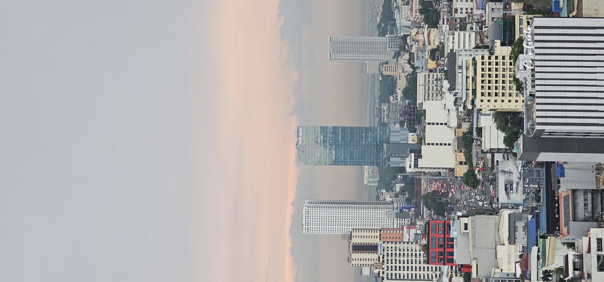 This is a panoramic, high-angle shot of the Makati City skyline in the Philippines, captured during twilight. The sky displays a gradient of soft pink and pale blue hues, with scattered, hazy clouds. In the foreground and mid-ground, a dense urban landscape unfolds, characterized by a mix of contemporary high-rise buildings, mid-rise structures, and lower-rise buildings.
Dominating the center of the frame is the Trium Square building, a modern skyscraper with a glass facade that reflects the sky. To its left stands a tall, white residential building with a grid-like pattern of windows. Further to the right, another white skyscraper is visible. Below these prominent towers, the city is a tapestry of various architectural styles and colors, with buildings ranging from cream-colored facades to bright red structures.
A busy, multi-lane road is visible in the lower mid-ground, filled with traffic, evidenced by the glowing red tail lights of vehicles and the headlights of oncoming cars, suggesting rush hour. Billboards are interspersed among the buildings. Some trees and patches of green are visible, offering glimpses of nature within the urban expanse. The overall impression is of a vibrant, bustling metropolis under a serene evening sky.