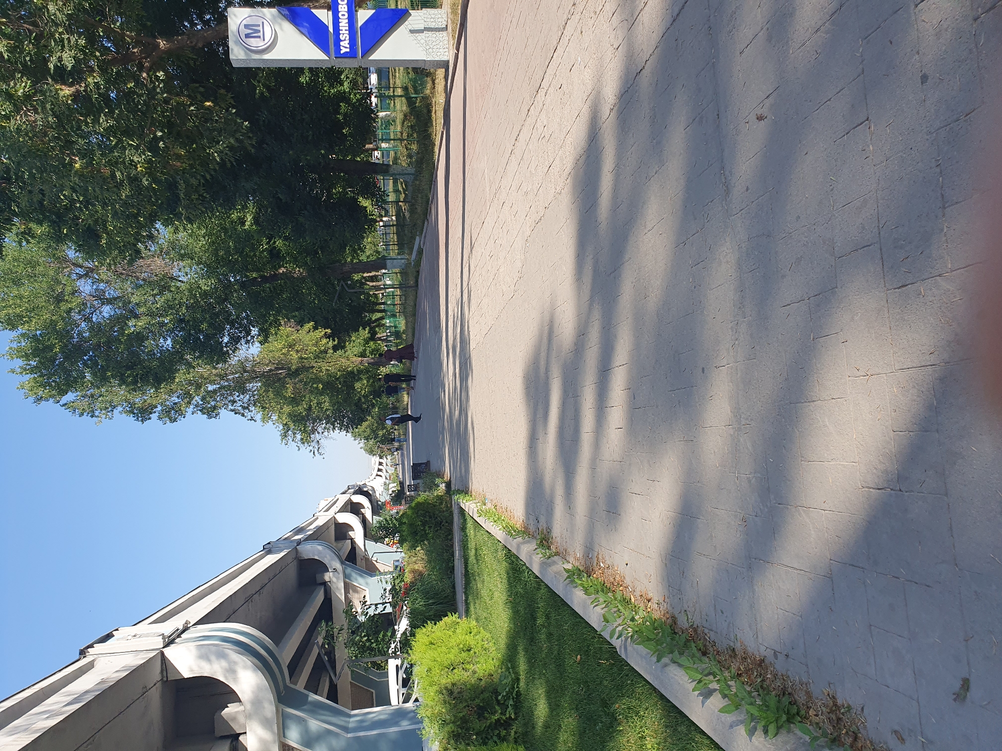 The image depicts a street scene in Tashkent, Uzbekistan, specifically near the Yashnobod metro station. The foreground is dominated by a paved walkway with a pattern of rectangular stones. To the left, a well-maintained green lawn with a concrete border transitions into a grassy area with some bushes. Beyond the grass and bushes, the underside of an elevated highway or overpass is visible, supported by concrete pillars. The highway structure is painted in white and blue.
Further down the walkway, several people are walking, suggesting a public space with pedestrian traffic. Trees with lush green foliage line the right side of the walkway, casting shadows on the pavement. A blue and white sign with a white "M" inside a circle and the word "YASHNOBOD" in blue letters is visible on the right side of the image, indicating the location of the metro station. In the background, a green metal fence encloses a grassy area, and more trees and buildings can be seen in the distance. The sky is clear and blue, indicating a sunny day.