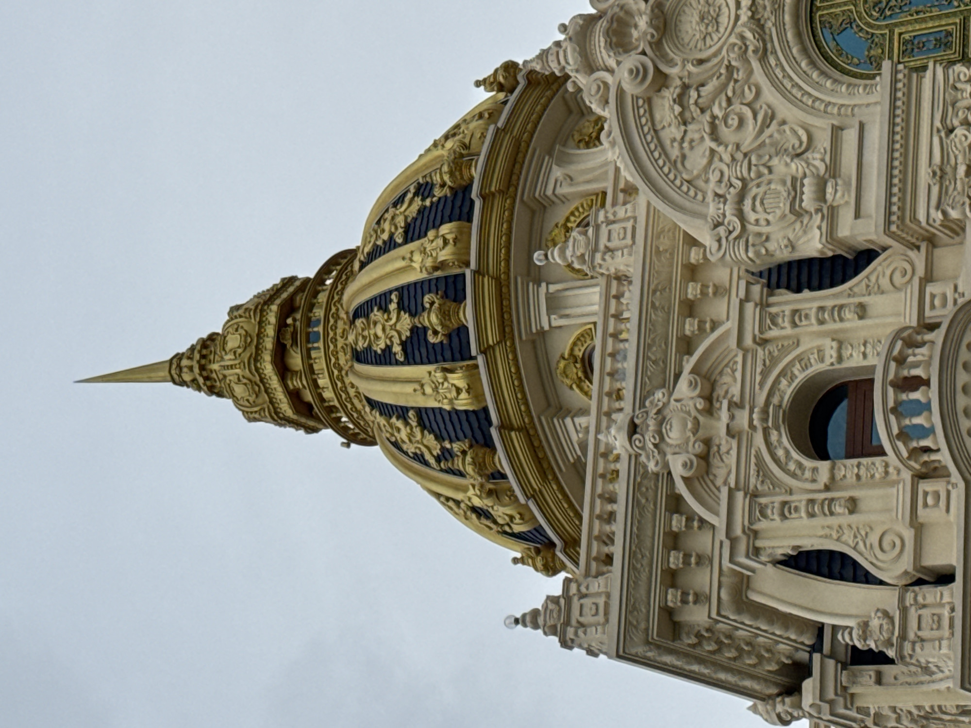 This image shows a close-up view of the golden dome and ornate facade of a building, likely a palace or a grand public building, in Da Lat, Vietnam. The dome is a striking feature, adorned with dark blue panels and intricate gold ornamentation, culminating in a tall, sharp spire. The building's exterior is characterized by elaborate stucco work, featuring decorative carvings, columns, and arched windows. The architecture suggests a European influence, possibly Beaux-Arts or French colonial style, common in Da Lat due to its history as a French hill station. The sky in the background is overcast and pale gray, providing a neutral backdrop that emphasizes the richness of the building's details. No people are visible in the frame.
