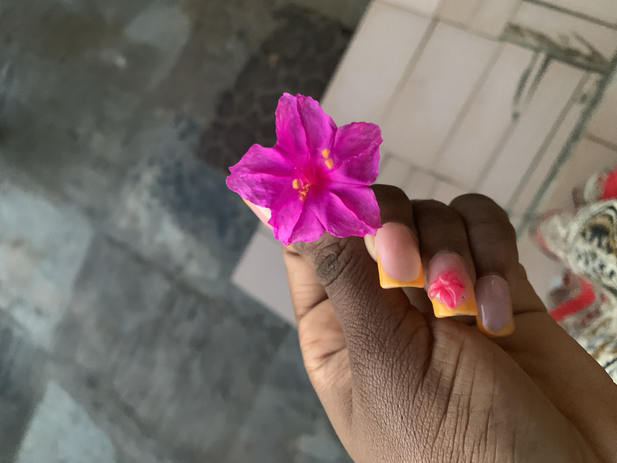 A hand with dark skin and brightly colored nails holds a vibrant pink flower. The flower, with its five petals and yellow stamen, is the central focus of the image. The nails are painted with a pink and orange gradient, and some have tiny pink floral designs. The background is blurred, showing a grey, tiled surface and a hint of a patterned fabric. The scene appears to be indoors, possibly on a floor or a table. There are no discernible city landmarks or specific location details.