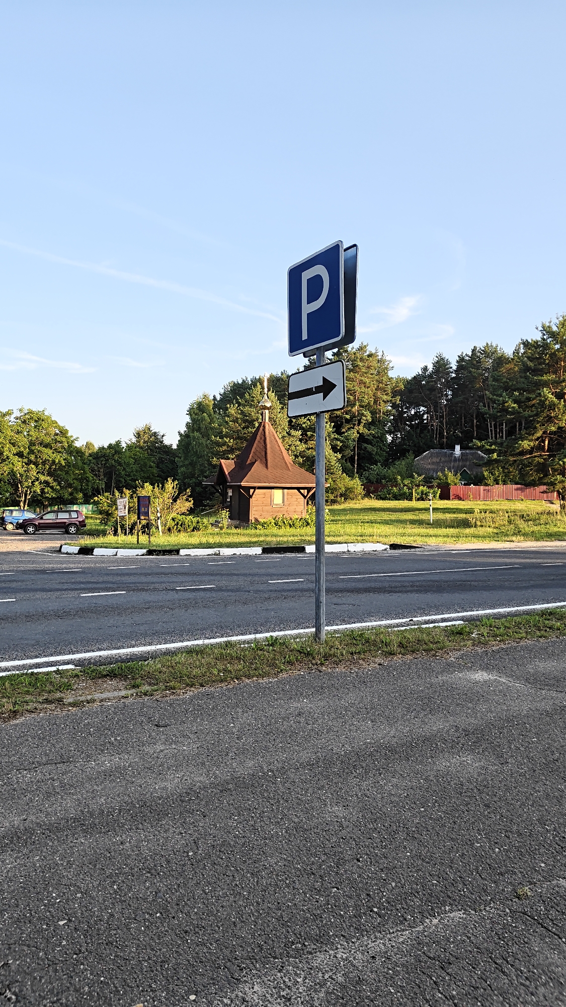 The image captures a view of a road and its surroundings on a sunny day in the village of Нарацкі сельскі Савет, Belarus. A blue and white parking sign with a white arrow pointing to the right is prominently displayed on a metal pole in the foreground. The road, paved with asphalt and bordered by white lines and a strip of grass, runs horizontally across the lower portion of the frame.
In the middle ground, behind the road, a small, traditional wooden building with a dark brown, steep, conical roof and a golden cross on top is visible. Adjacent to this building, a few cars are parked on a grassy area. Further in the background, a dense line of pine trees forms a backdrop, and a red wooden fence can be seen. The sky above is a clear, light blue with a few wispy clouds. The scene depicts a typical rural or semi-rural setting, likely a point of interest or amenity for visitors to the area.