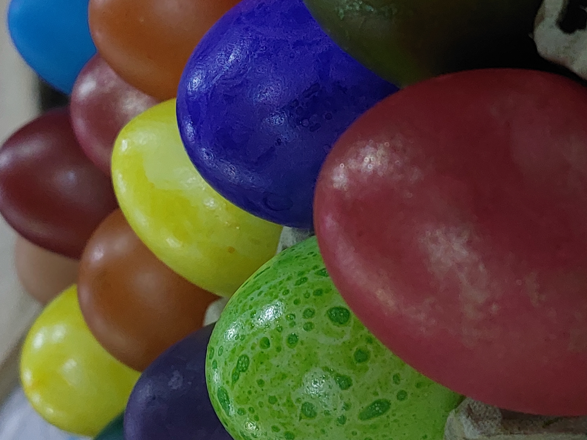 The image shows a close-up of a collection of multicolored, round candies, likely chocolate-covered. The candies are arranged haphazardly, overlapping each other. Colors present include blue, brown, red, yellow, green, and purple. The green candy has small, darker green spots. The candies appear to have a slightly textured surface. The background is blurry and out of focus. The location is unknown.