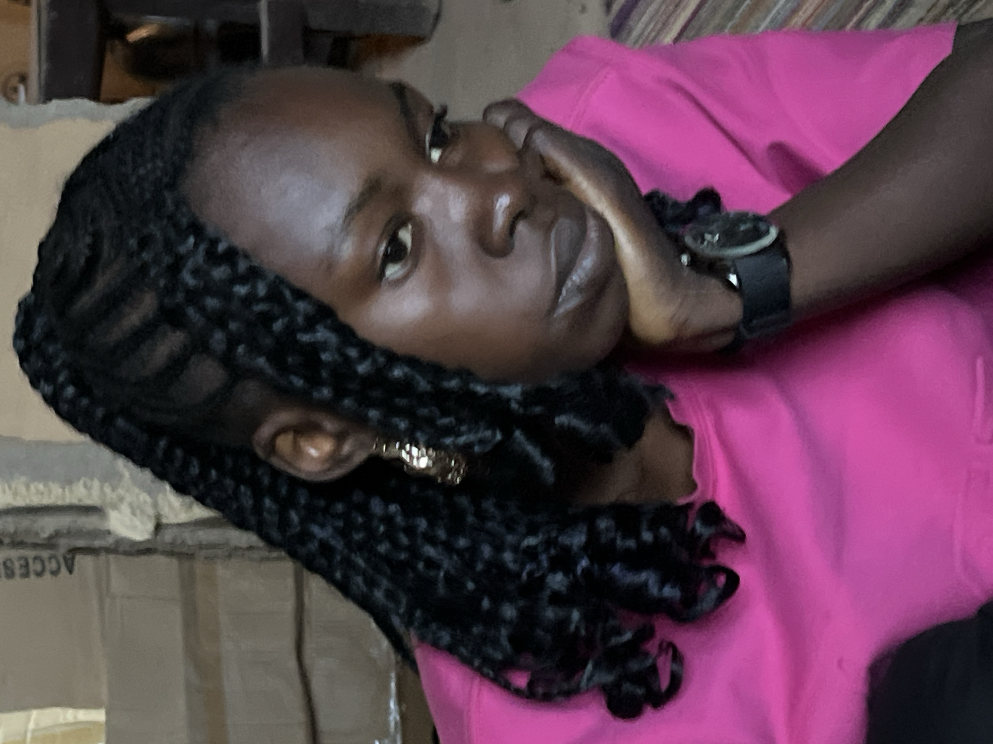 A close-up shot captures a young Black woman with dark skin, wearing a bright pink hoodie, her face tilted downward and resting on her right hand. Her hair is styled in braids with loose, curly ends. She wears a large, gold earring in her visible left ear and a black watch on her right wrist. The background is indistinct, with hints of cardboard boxes and muted tones suggesting an indoor setting. The image is shot from a slightly low angle, emphasizing the woman's thoughtful or somber expression. The lighting highlights the sheen on her skin, particularly on her cheek and forehead.