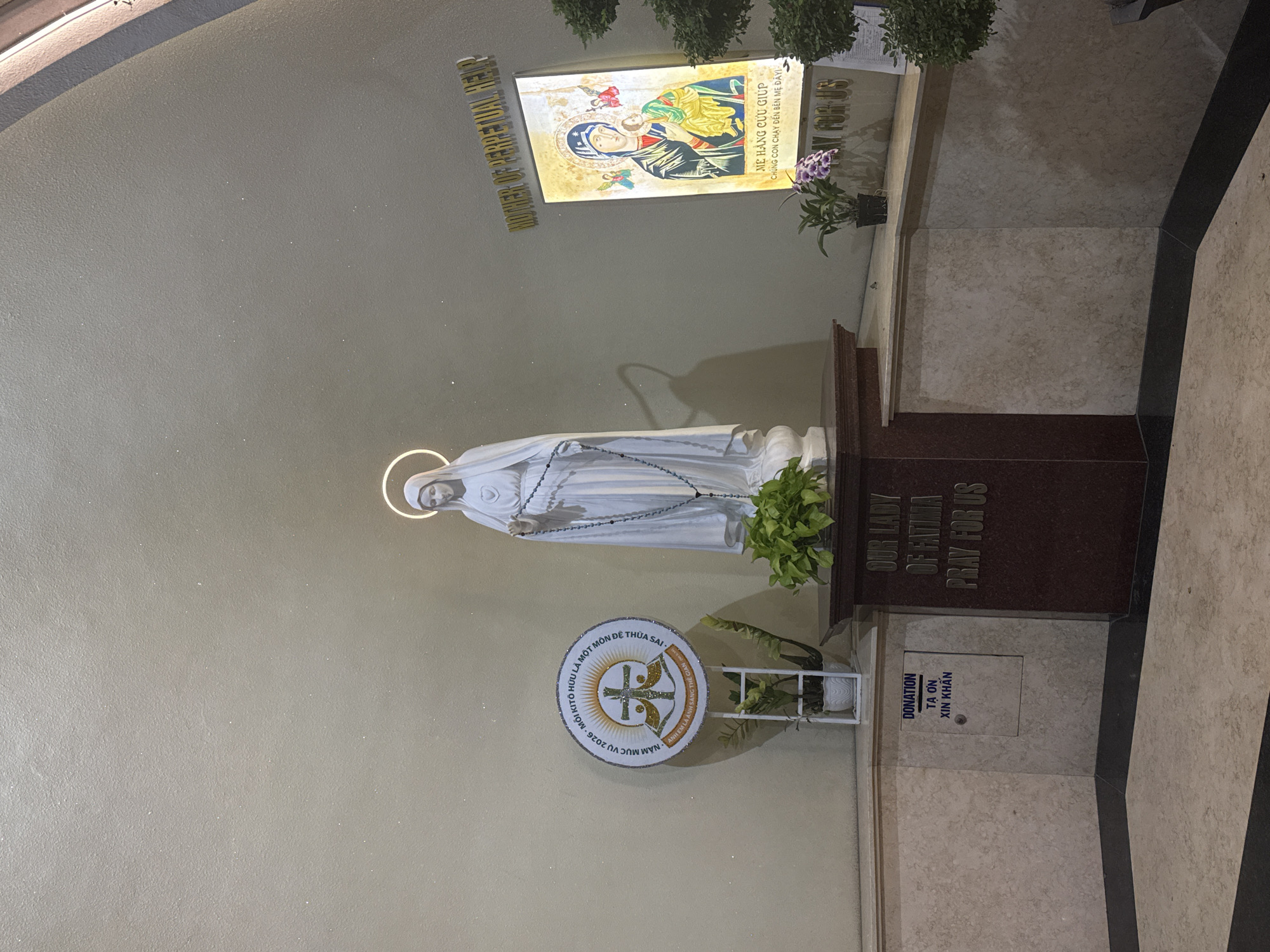 The image depicts an interior view within a religious setting, specifically a church or chapel. Dominating the scene is a statue of the Virgin Mary, wearing a white robe and holding a rosary, positioned on a brown marble stand inscribed with "OUR LADY OF FATIMA PRAY FOR US." To the left of the statue is a round sign with Vietnamese text and a religious symbol, while a display with the words "MOTHER OF PERPETUAL HELP" is placed higher up. A donation box labeled "DONATION TẠ ƠN XIN KHẨN" is seen below the statue's stand. The setting appears to be in Nha Trang Ward, Vietnam.
