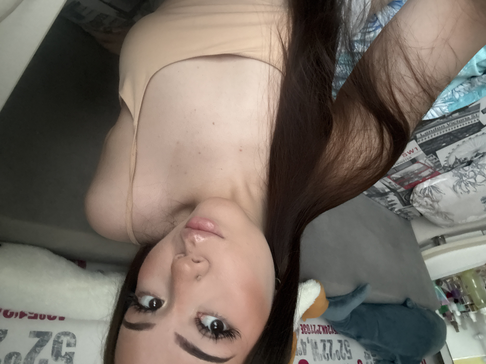 A rotated close-up image shows a woman with dark hair, fair skin, and noticeable makeup, including arched eyebrows and heavy mascara. She is wearing a light beige tank top. In the background, various objects are visible, including a grey stuffed whale and a pillow. There's also a newspaper with the text "London Victoria St SW1" visible, which suggests a setting unrelated to Naberezhnye Chelny, Russia. The location information appears to be part of the newspaper print or other background material in the image.