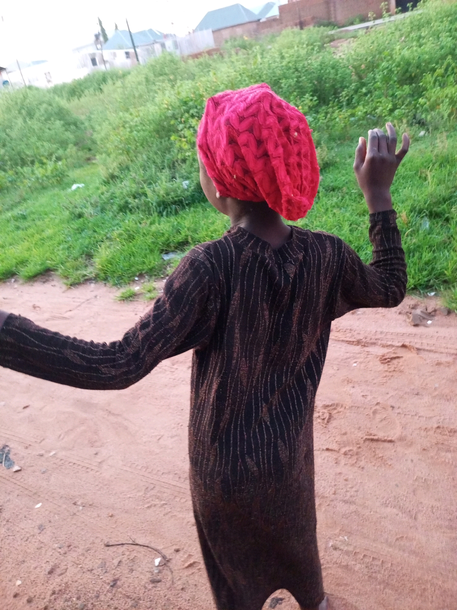 A person wearing a dark, long-sleeved garment with a pattern of thin, vertical, metallic-colored stripes and a bright red headscarf is standing outdoors on a dirt path. Their back is to the camera, and their head is turned to the left, with their left arm raised and hand extended, palm facing forward, fingers slightly curled. To the left of the person, there is a patch of dry, sandy ground with some debris. Behind the person, a grassy slope rises, dotted with green vegetation and some white debris. In the distance, several buildings with light-colored walls and slanted roofs are visible. The sky is overcast. This scene is consistent with an outdoor environment in the city of Tempure, Nigeria.