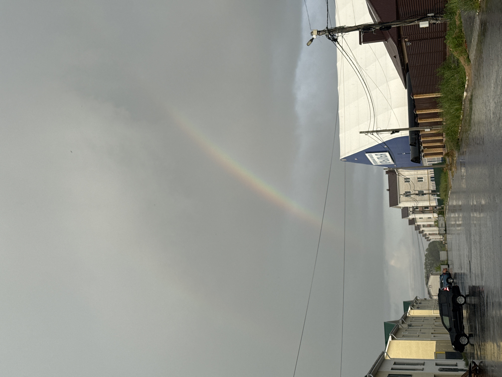 A double rainbow arcs across a cloudy, gray sky over a street in Murom, Russia, after a rain shower. The street is wet and reflective. On the left, several two-story residential buildings with yellow and white facades and green roofs line the street. A black SUV is parked on the left side of the street, and another blue car is visible further down. On the right side of the street, a large building with a white, tent-like roof and a blue section with Cyrillic text that reads "КРЫЛЬЯ" (Wings) is visible. Adjacent to it is a brown wooden fence and a street light on a wooden pole with numerous electrical wires. The overall impression is of a quiet, post-rain suburban or commercial area.