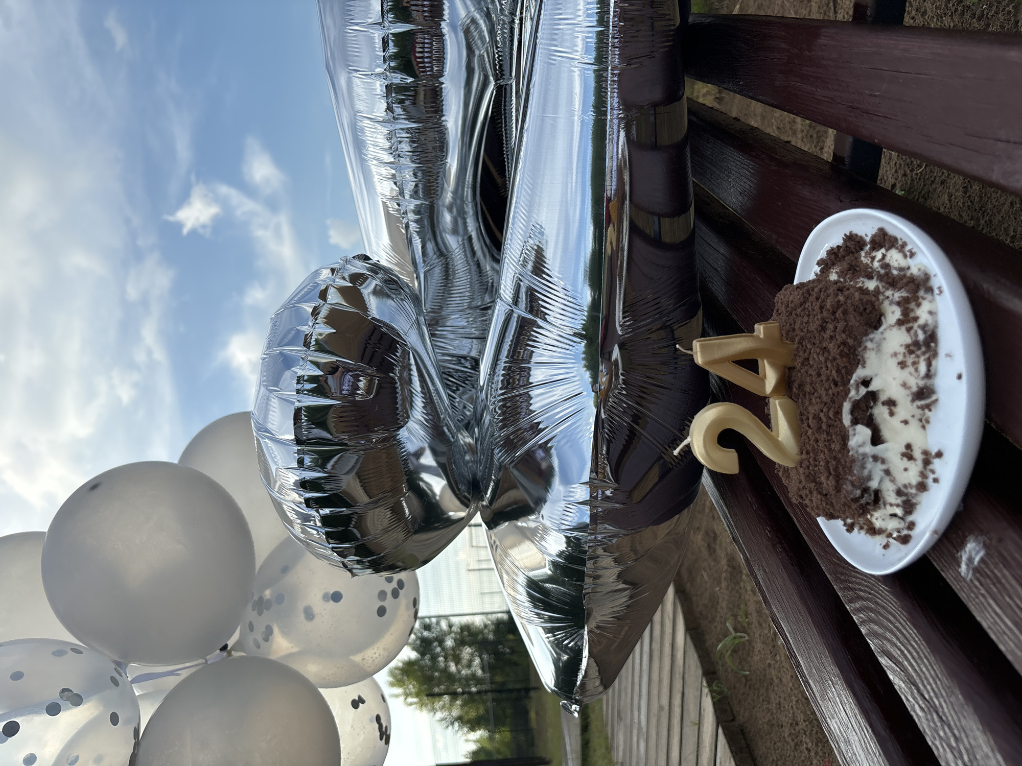 This image shows a chocolate cake with cream frosting on a white plate, decorated with two gold candles shaped as the numbers "2" and "4", indicating the age twenty-four. The cake is placed on a wooden bench, with a portion of a dark brown wooden surface visible in the foreground. Behind the cake, a cluster of silver and white balloons, some filled with confetti, are visible, obscuring much of the background. The balloons are reflective, mirroring the sky and surrounding trees. In the distance, a fence and trees are visible. The scene appears to be outdoors, likely in a park or backyard setting. The sky is blue with scattered white clouds. There are no people visible in the image. The image does not contain any information that can be used to determine the location in the city of Chaikovsky, Russia.