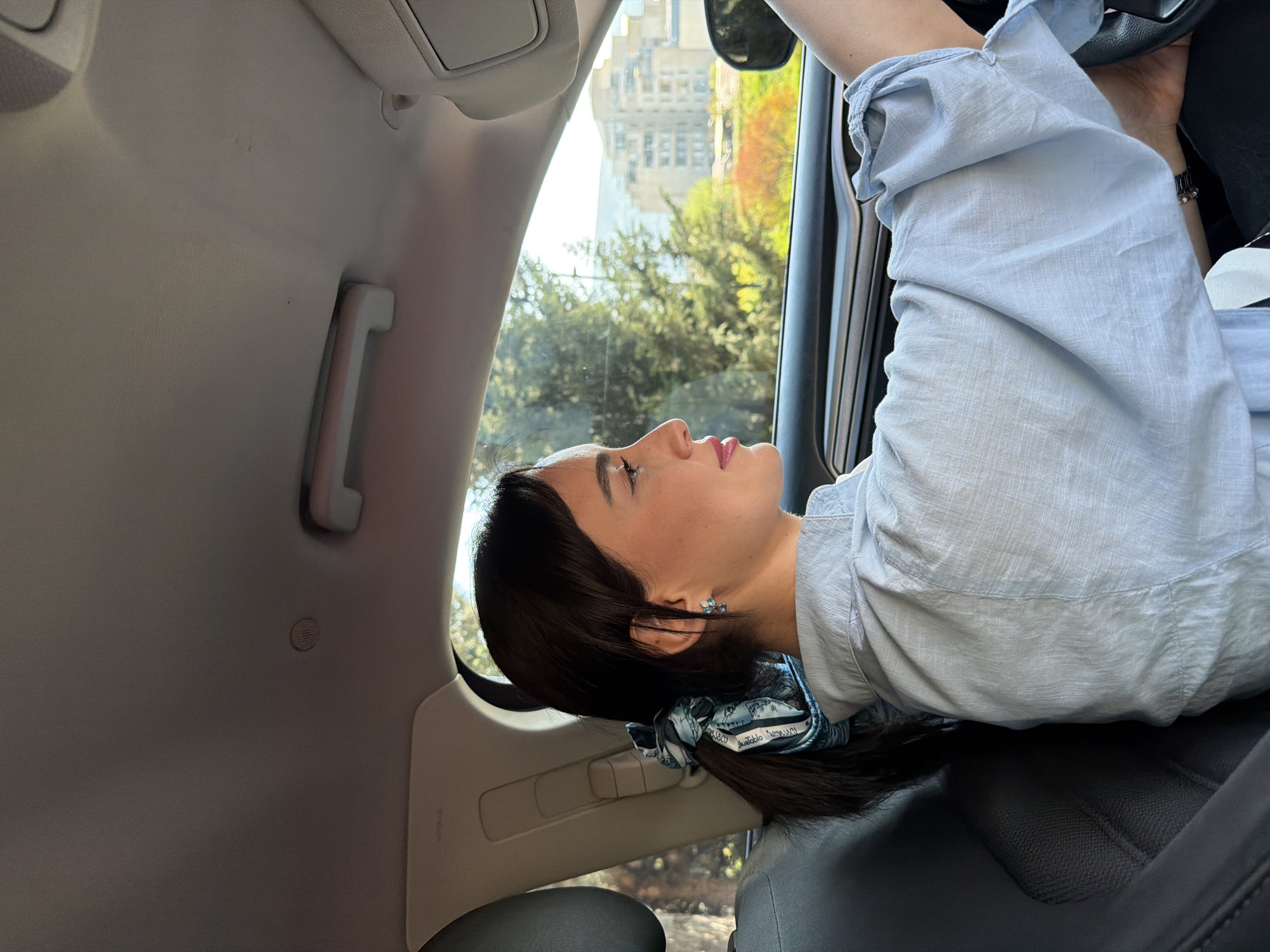 The image is rotated 90 degrees clockwise, depicting a young woman from a side profile, leaning back or resting inside a car. The car's headliner is visible on the left side of the frame, with a light-colored grab handle.
The woman has dark hair pulled back, secured by a blue and white patterned scrunchie. She wears a light blue, long-sleeved shirt. A small blue butterfly-shaped earring adorns her visible ear. Her lips are a soft pink.
Through the car window, illuminated by bright daylight, an urban landscape is visible. Green trees with autumn foliage hints and a large, light-colored building with numerous windows can be seen, consistent with a city environment like Tbilisi, Georgia.
Inside the car, a dark car seat is prominent beneath her. Parts of the car's interior paneling and what appears to be a steering wheel or dashboard are also visible. A small circular vent or speaker is embedded in the headliner. On the scrunchie, faint text can be seen, with parts like "olablo" and "BUSCO" discernible.