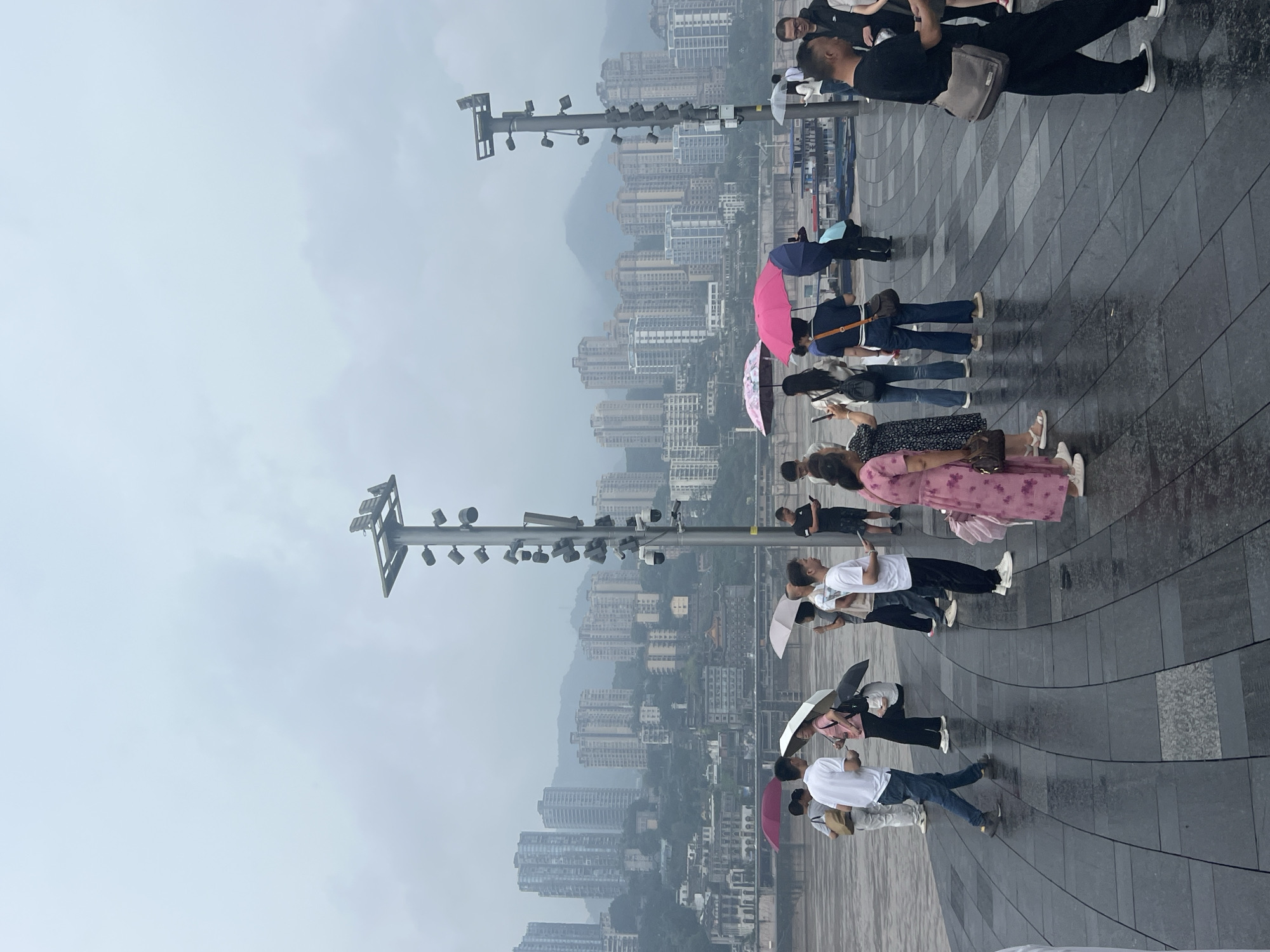 A group of people are standing on a wet, tiled promenade in front of a river, with a city skyline of multi-story buildings and mountains in the background. It appears to be a cloudy or overcast day. Several individuals are holding umbrellas, suggesting it is raining or has recently rained. The people are casually dressed, and some are interacting with each other. There are also lampposts with multiple lights and security cameras visible along the promenade. The architecture of the city suggests an urban environment, possibly Yuzhong District, China, known for its hilly terrain and riverside development.