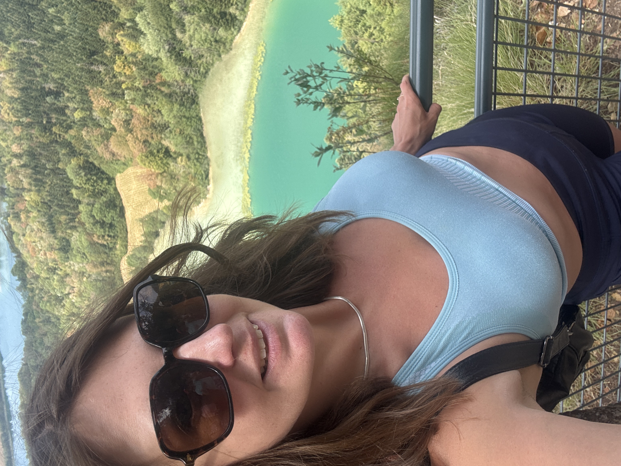 In this selfie, a woman with brown hair and sunglasses stands on a viewing platform, leaning on a dark green metal railing. She is wearing a light blue sports bra and dark blue shorts. Behind her, a vibrant turquoise lake is visible, surrounded by lush green trees and hills. The scene appears to be a scenic overlook, possibly at Lac d'Ilay, a natural lake in the Jura region of France, near the commune of La Chaux-du-Dombief. The lighting suggests it is daytime.