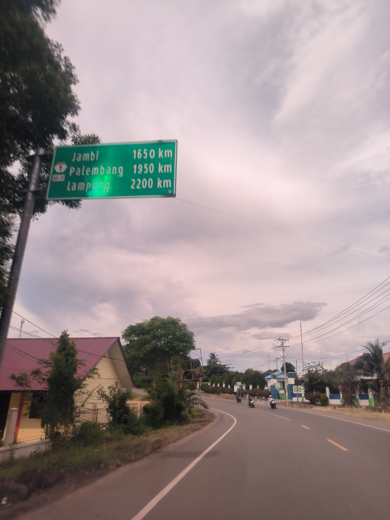 This full-shot image captures a view from a moving vehicle on an asphalt road in Lampanahtunong, Indonesia, under an overcast sky. The road features yellow double solid lines in the center and a white solid line on the right shoulder, curving slightly into the distance.
On the left side of the road, a prominent green overhead road sign is visible. The sign, supported by a pole, displays destinations and distances in white text: "Jambi 1650 km", "Palembang 1950 km", and "Lampung 2200 km". Below "Jambi", there's a white shield icon with a red top and the number "1", along with smaller text "AH25". Further down the left, there's a building with a maroon corrugated roof and light-colored walls, partially obscured by lush green trees and foliage.
On the right side of the road, several motorcycles with riders are seen traveling in the same direction. Beyond them, a line of buildings with blue and white accents, surrounded by trees and utility poles with overhead power lines, stretches along the roadside.
The sky is cloudy, exhibiting light grey and soft pinkish hues, suggesting either early morning or late afternoon light. The overall scene depicts a functional road infrastructure in a tropical or subtropical region, with long-distance travel information provided.