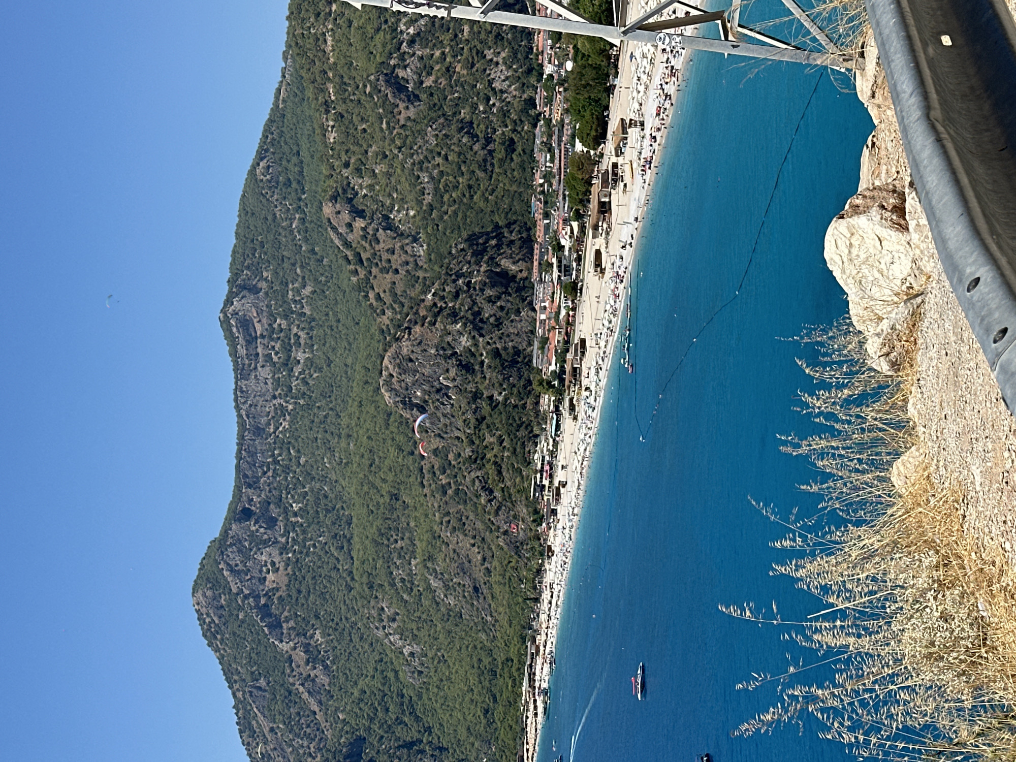 A high-angle, wide shot captures a vibrant coastal scene under a clear, bright blue sky. In the foreground, dry, tan-colored grasses grow on a rocky incline, with a metal guardrail running horizontally across the bottom of the frame. Beyond the grasses, a vast expanse of intensely blue, calm water stretches out, dotted with a few small boats.
A long, white-sand beach lines the shore, densely populated with numerous sunbathers lounging on white beach chairs and umbrellas. A strip of buildings, a mix of low-rise structures with red and white roofs, forms a town behind the beach. The backdrop is dominated by a large, green, tree-covered mountain with rocky, barren peaks. Two paragliders are visible in the sky above the mountain. The scene appears to be a popular tourist destination during daylight hours.