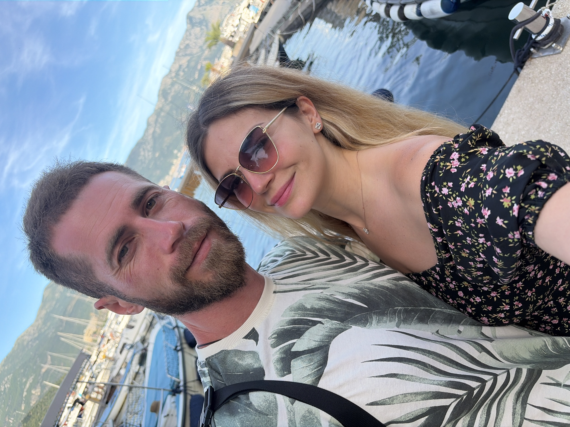 A man and a woman are taking a selfie in Budva, Montenegro. The man, with a beard and short brown hair, is wearing a white t-shirt with a green palm leaf print. The woman, with blonde hair, is wearing sunglasses and a black off-the-shoulder dress with a floral pattern. They are standing by a body of water, with boats docked in the background. Mountains are visible in the distance. The scene appears to be in a marina.