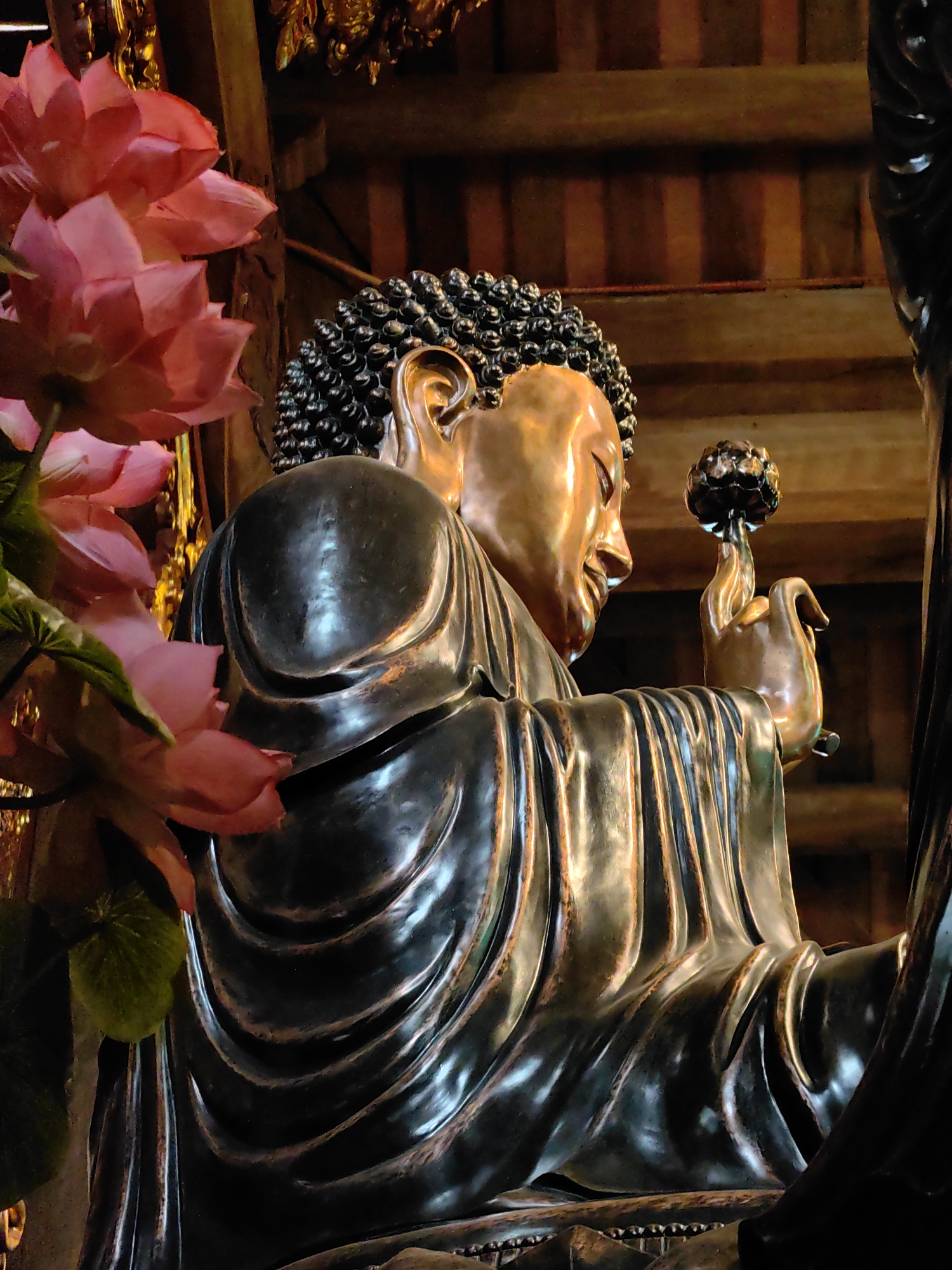 A statue of Buddha is positioned in Hong Gai Ward, Vietnam. The bronze-colored figure is holding a dark lotus flower bud. The head features tight, dark curls. A large arrangement of pink lotus flowers is placed next to the statue. The backdrop is wooden and slightly out of focus.