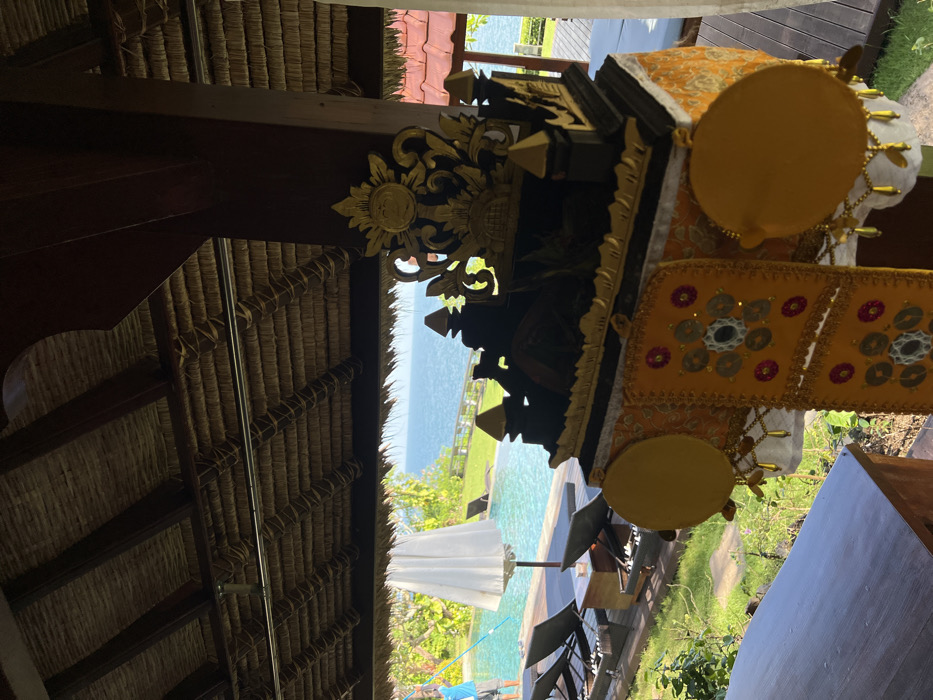The image depicts an outdoor tropical resort scene in Uluwatu, Indonesia, viewed from beneath a shaded structure with a thatched roof. In the immediate foreground on the left, a dark wooden surface, possibly a table or counter, is partially visible. Centrally, a Balinese ceremonial object, likely a shrine or offering stand, is prominent. This object is dark-colored with intricate gold carvings, featuring stylized pagoda-like structures and decorative flourishes, including a sun-like emblem at its peak. It is draped in patterned yellow and orange fabric, adorned with multiple circular yellow discs and smaller, coin-like reflective embellishments arranged in geometric patterns. The overhead view reveals a densely woven thatched roof supported by dark wooden beams.
In the midground, a light blue infinity pool is visible, surrounded by a dark wooden deck. Several dark-colored lounge chairs with white cushions are arranged on the deck. A large, open white umbrella provides shade near the pool. To the left, a single individual wearing a light blue shirt is partially visible, actively engaged in cleaning the pool using a long pole.
The background features an expansive view of the blue ocean extending to the horizon under natural light. A manicured green lawn separates the pool deck from the ocean, bordered by a white railing. Distant lush green trees and foliage line the coastline, and a structure with red-tiled roofs is partially observable on the far right.