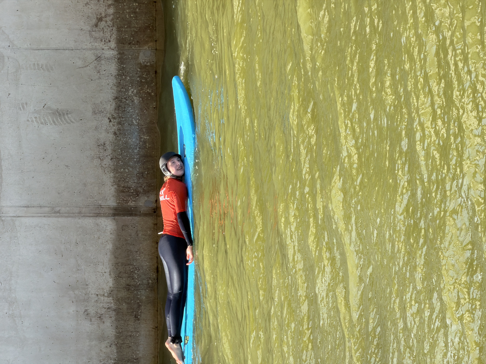 A person wearing a red long-sleeved shirt, black leggings, and a helmet is lying face down on a bright blue surfboard in murky yellow water. The person's head is turned to the left, and they are smiling. The surfboard is floating on the water, which appears to be in an urban setting. In the background, there is a large concrete wall with visible texture and some staining. The scene is set in the Surf Center Rotterdam, an artificial wave pool located in the port of Rotterdam, Netherlands. The pool is designed to generate waves for surfing, and the concrete wall likely forms part of the pool's structure or the surrounding urban landscape. The water is a characteristic greenish-yellow color due to the mineral content and algae growth common in artificial wave pools. The lighting suggests it is daytime, with sunlight reflecting off the water's surface, creating ripples and highlights.