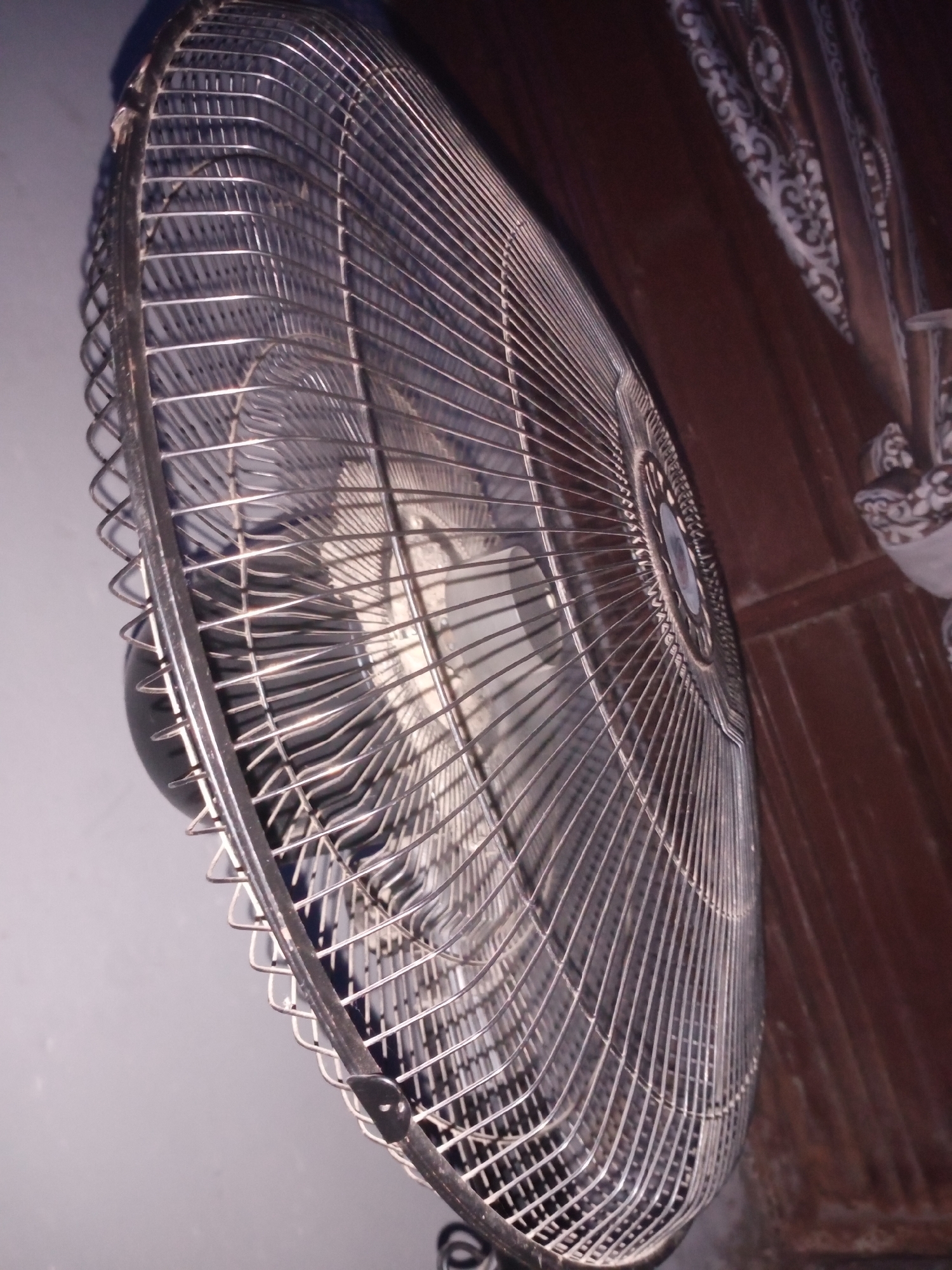The image is a close-up, low-angle shot of a metal fan grill. The fan is positioned in a way that the grill dominates the frame, with its intricate metalwork casting shadows. The background is divided into two distinct areas. To the left, a plain, light-colored wall is visible. To the right, a darker, textured surface, possibly a wooden door or paneling, is present, adorned with decorative patterns. The lighting is dim, with a strong highlight on the fan's metal. No people or actions are depicted. The scene appears to be indoors, possibly in a residential or commercial setting in Jalingo, Nigeria.