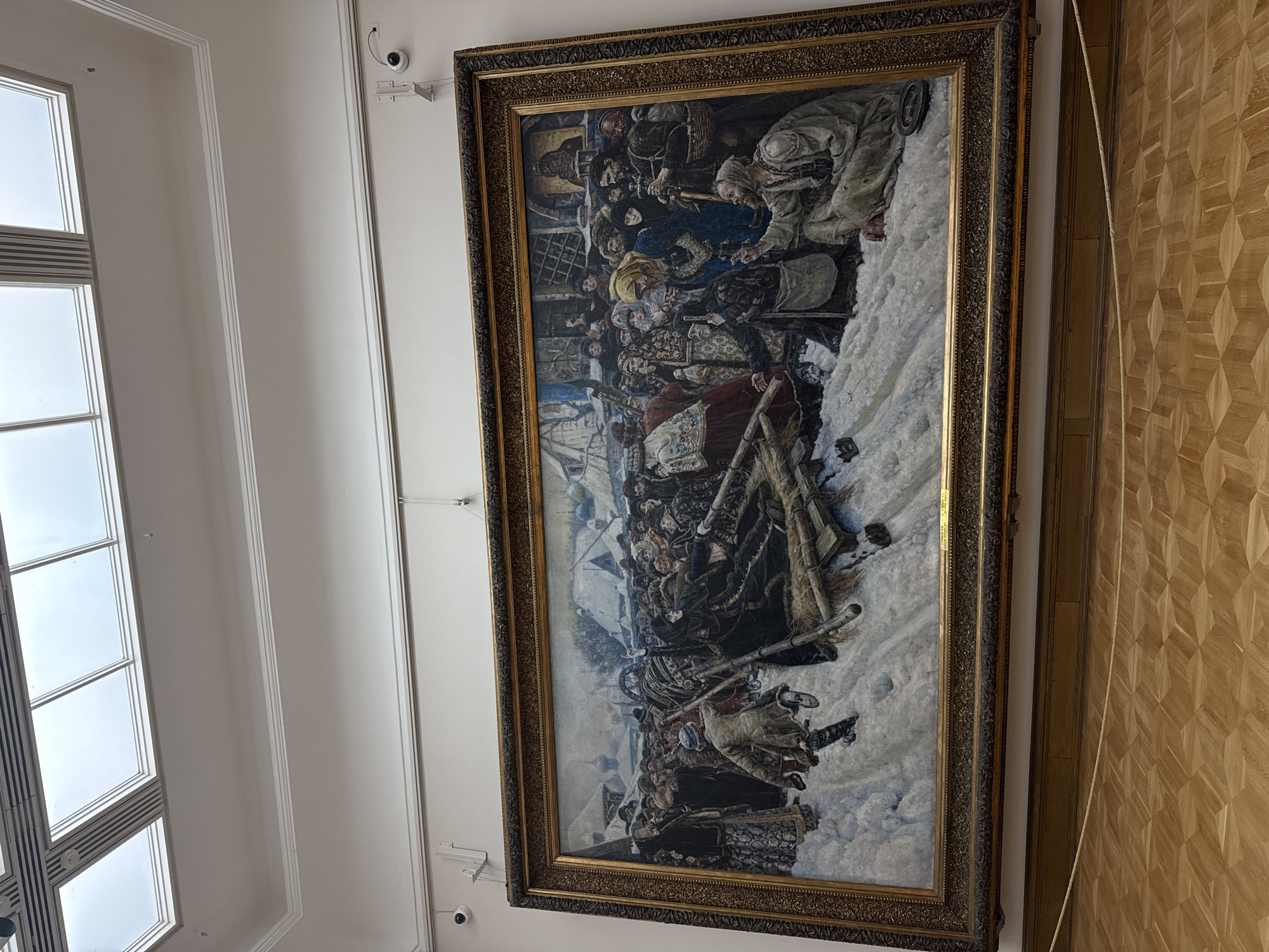 A large, ornately framed painting hangs on a wall in what appears to be a building with herringbone pattern wood flooring. The painting depicts a large group of people in a snowy, historical setting. Some individuals are dressed in traditional clothing and appear to be involved in a battle or siege. The setting resembles an outdoor winter scene, with details of people, weapons, and snow-covered ground. There is a window to the left of the painting. The security cameras are placed above the painting. It is possible that the painting is displayed at the State Tretyakov Gallery in Moscow, Russia.