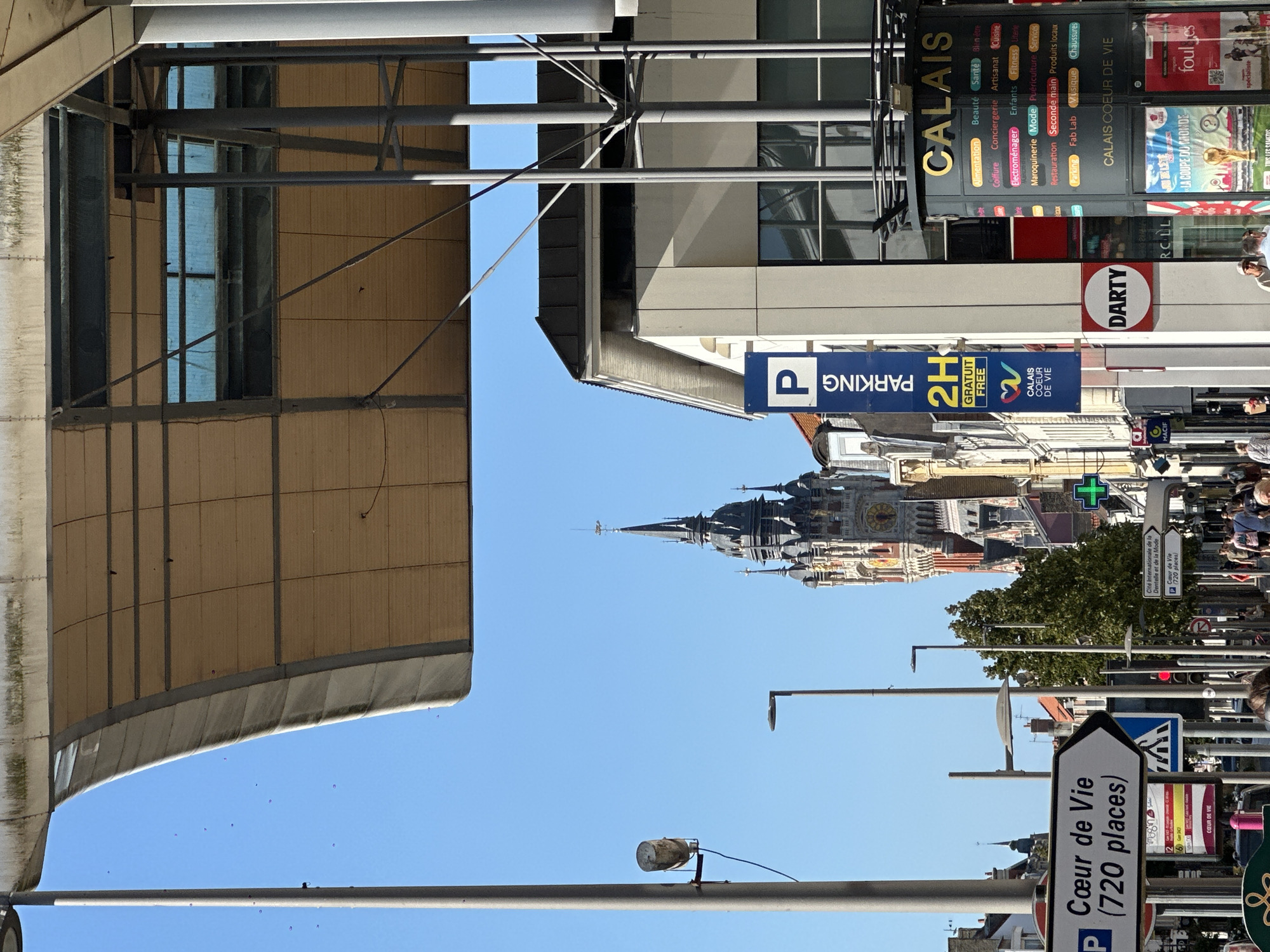 The image depicts a street scene in Calais, France, centered around a tall, ornate clock tower. A modern canopy structure with a beige, perforated ceiling juts out from the top of the frame, supported by gray metal beams and pillars. Below the canopy, the clock tower, with its distinctive spires and clock face, stands prominently against a clear blue sky. To the right of the tower, a white building with a dark sloped roof is visible, along with a vertical blue sign for "PARKING 2H GRATUIT FREE" and another multi-colored circular sign for "CALAIS COEUR DE VIE" listing various shops and services.
On the left side of the street, a white sign with a black arrow points to the right, indicating "Cœur de Vie (720 places)". Further down the street, more buildings, streetlights, and trees line the sidewalk, where several people are walking. One person is wearing a blue top and black shorts, while others are dressed in casual attire. A green cross, likely for a pharmacy, is visible on the right side of the street. The overall environment appears to be an urban commercial district.