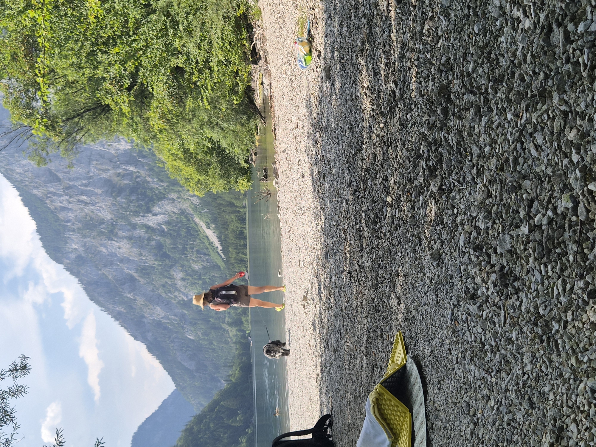 In Eisenerz, Austria, a person wearing a straw hat and a backpack stands on a pebble beach, holding a leash attached to a dog that is partially in the water. The person is wearing shorts and yellow shoes. Behind them, a calm lake is surrounded by lush green trees and imposing, rocky mountains under a bright, partly cloudy sky. In the foreground, a yellow and gray mat is laid out on the pebble-covered ground, and a black backpack is leaning against something on the left side of the frame. Further down the beach, a yellow and green inflatable object is visible.