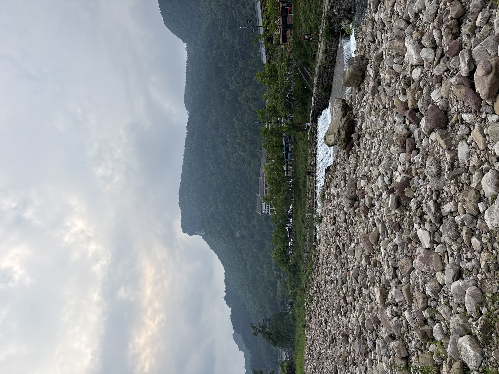 The photograph depicts a rocky landscape in Wulingyuan, China. A mountain range, covered in dense green vegetation, rises prominently in the background, partially obscured by a grey, overcast sky. At the base of the mountain, structures, including buildings and parked vehicles, are visible amongst trees. In the foreground, a stream cascades over a series of small weirs. The ground is covered in a dense layer of rounded rocks, with sparse vegetation.