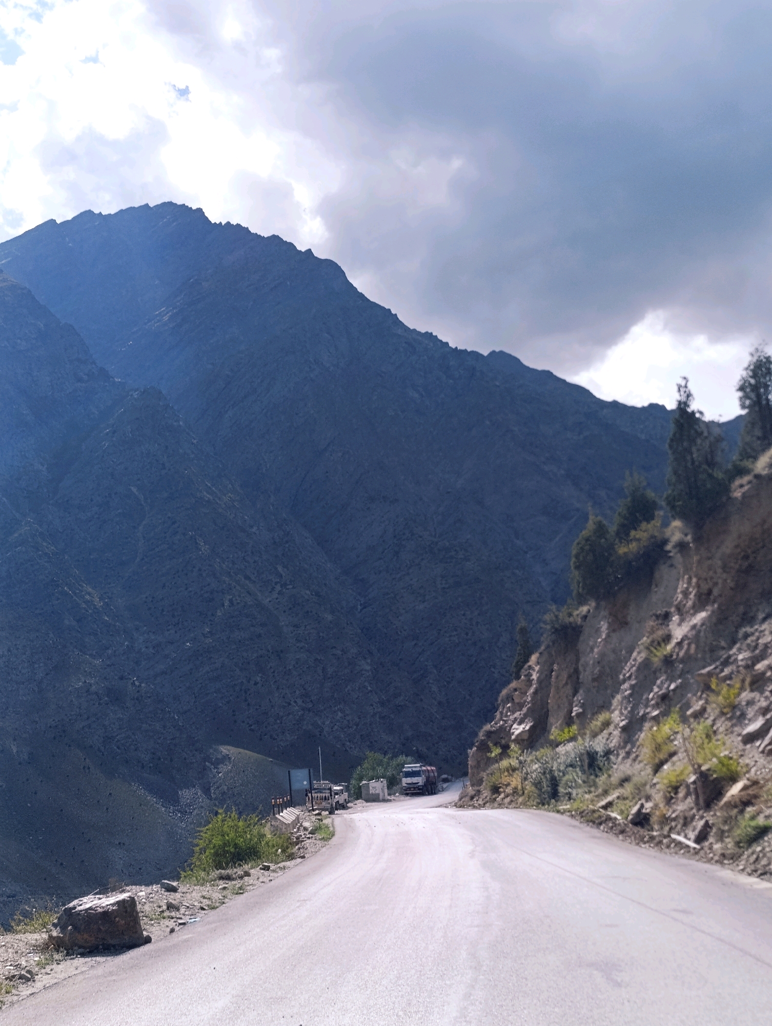 This is a landscape photograph taken from a road in Darcha, India. A paved road curves into the distance, bordered by a steep, rocky mountainside on the right, dotted with sparse vegetation and trees. On the left, a massive, dark-colored mountain range dominates the scene, with its rugged slopes stretching towards a cloudy sky. Several vehicles, including a truck and what appears to be a utility vehicle, are visible parked or driving along the road in the middle ground, near a small cluster of buildings and structures. The sky above is partly cloudy, with patches of bright sunlight breaking through.