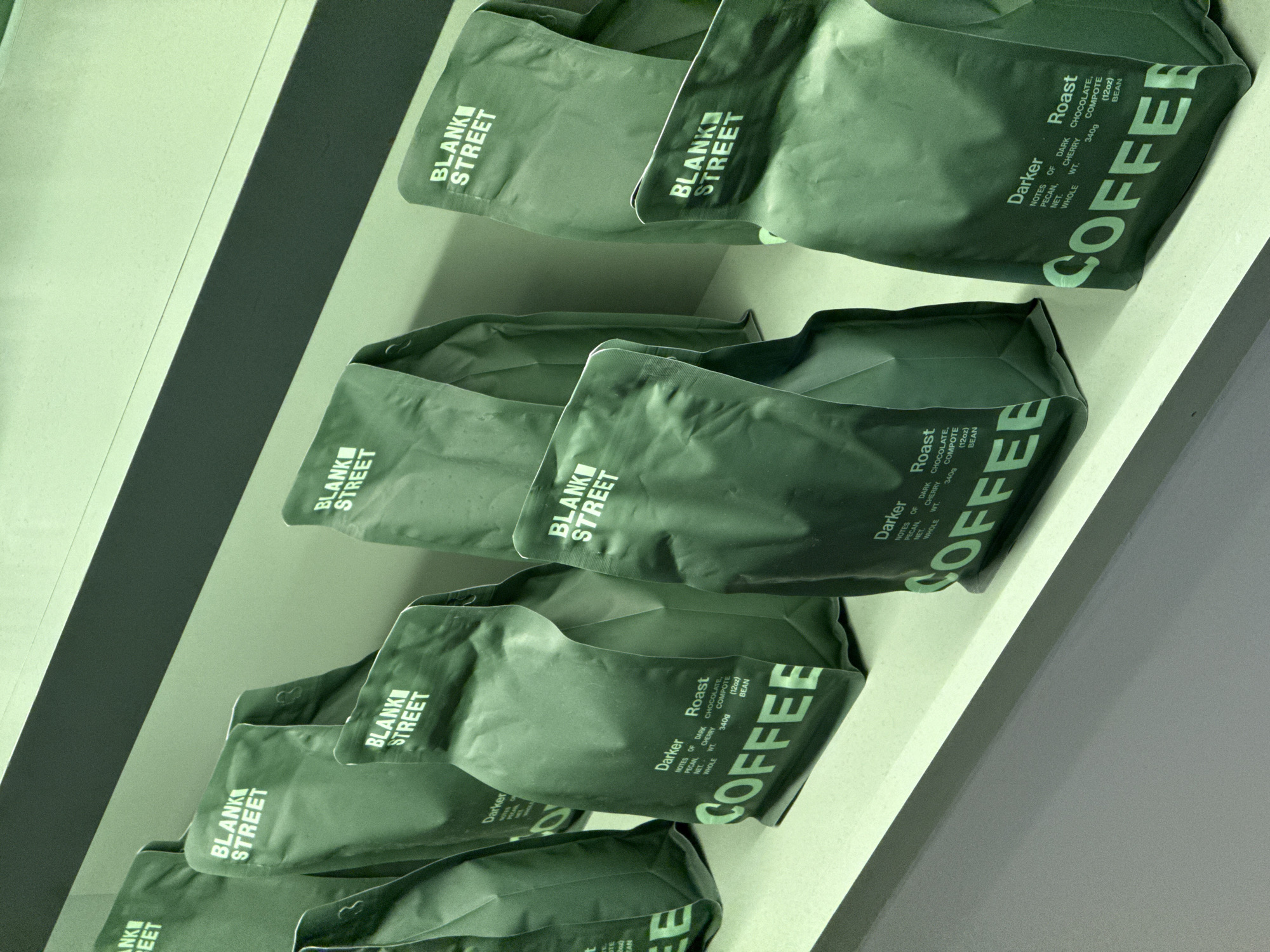 The image depicts several bags of coffee from "Blank Street" on a shelf. The bags are green and labeled with "Darker Roast", detailing notes of dark chocolate, pecan, and cherry, and specifying a net weight of 340g. The bags are arranged in rows, with the "Blank Street" logo visible on each. The setting appears to be a retail environment, possibly a café or store in Boston, United States.