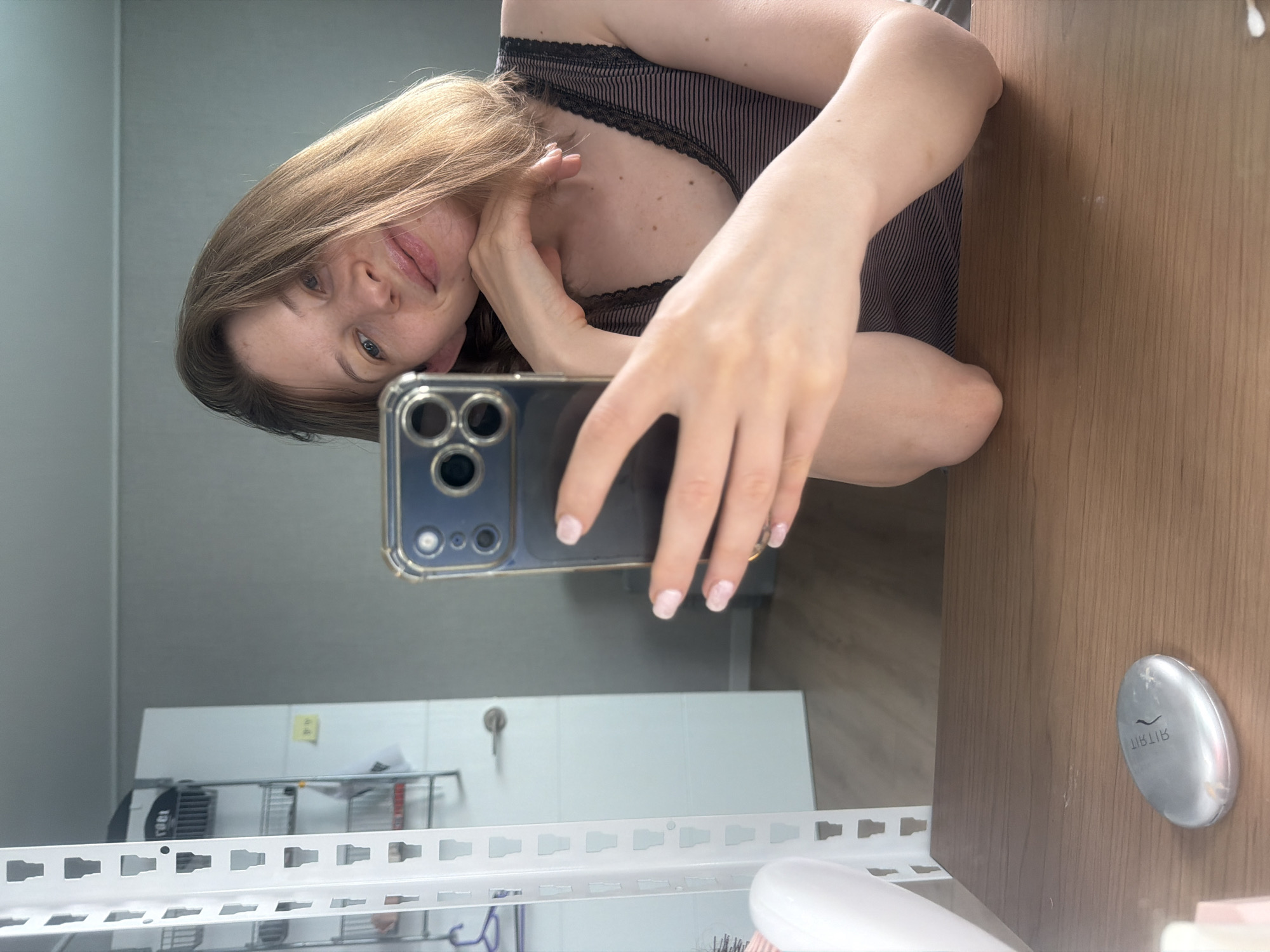A woman with blonde hair and light blue eyes is taking a selfie in a room. She is leaning her chin on her hand and looking at the camera. She is wearing a striped tank top. In front of her is a wooden table with a silver oval-shaped object. To her left, a white shelf unit is visible, with a white door and a rack of items behind it. The room appears to be a bedroom or dressing area, possibly in an apartment in Dongducheon-si, South Korea, given the general interior style.