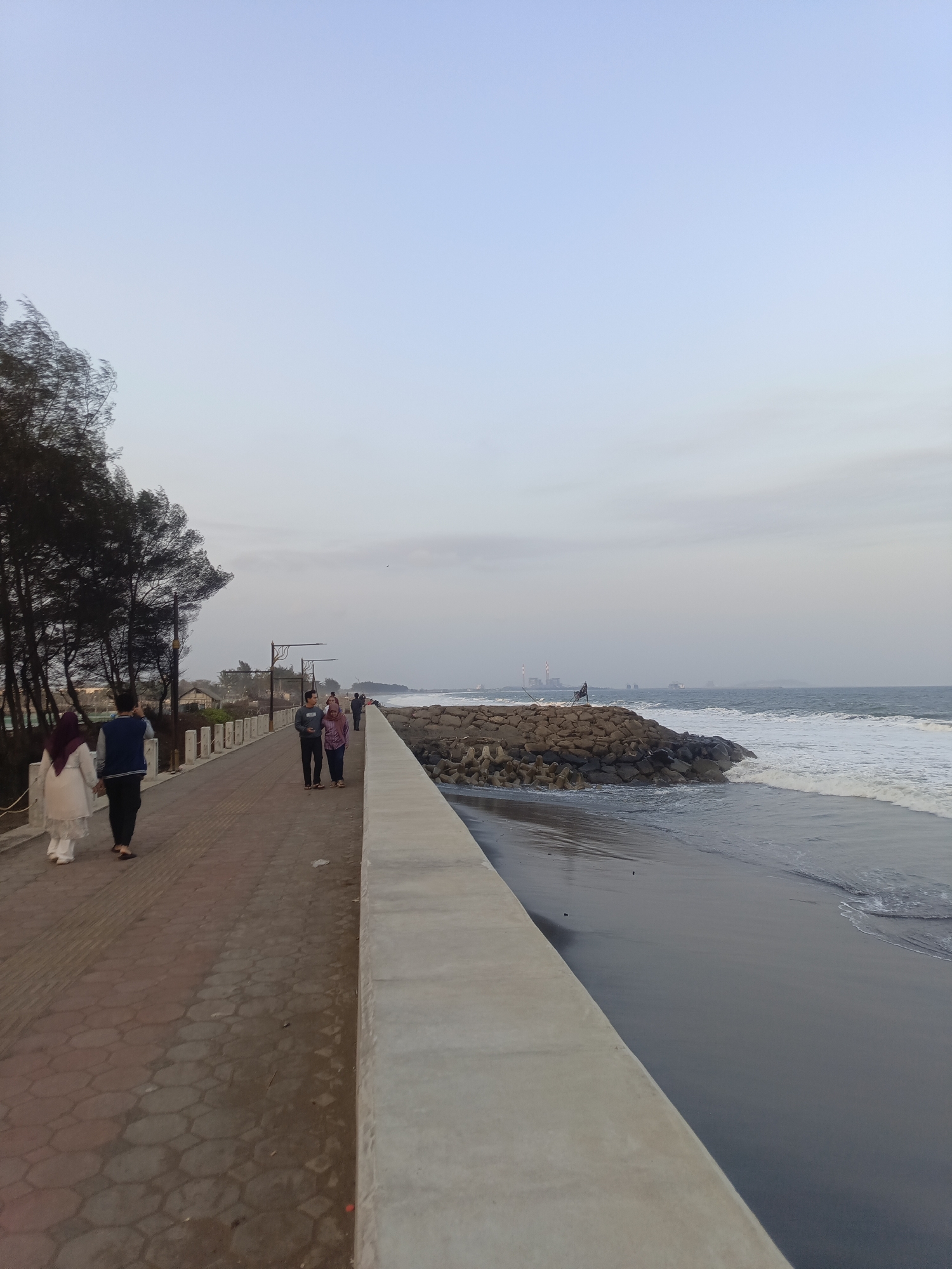 The image shows a promenade next to a beach in Kelurahan Tegalkamulyan, Cilacap, Indonesia. The sky is clear and a pale blue, with some clouds near the horizon. The sea is calm with moderate waves breaking on the dark sand beach. A concrete breakwater extends into the sea, constructed with large rocks and concrete blocks.
On the promenade, several people are walking. In the foreground, a couple is walking away from the camera, with the man wearing a blue and white jacket and dark pants, and the woman in a white hijab and dress. Further down the promenade, another couple is walking towards the camera. Behind them, more people can be seen walking.
To the left of the promenade, there is a line of trees with dark foliage, and beyond them, some buildings and structures are visible, including what appears to be a factory with tall chimneys in the distance. Streetlights with decorative tops are spaced along the promenade. The promenade itself is paved with hexagonal tiles and has a low concrete barrier. The overall scene suggests a public space used for recreation.