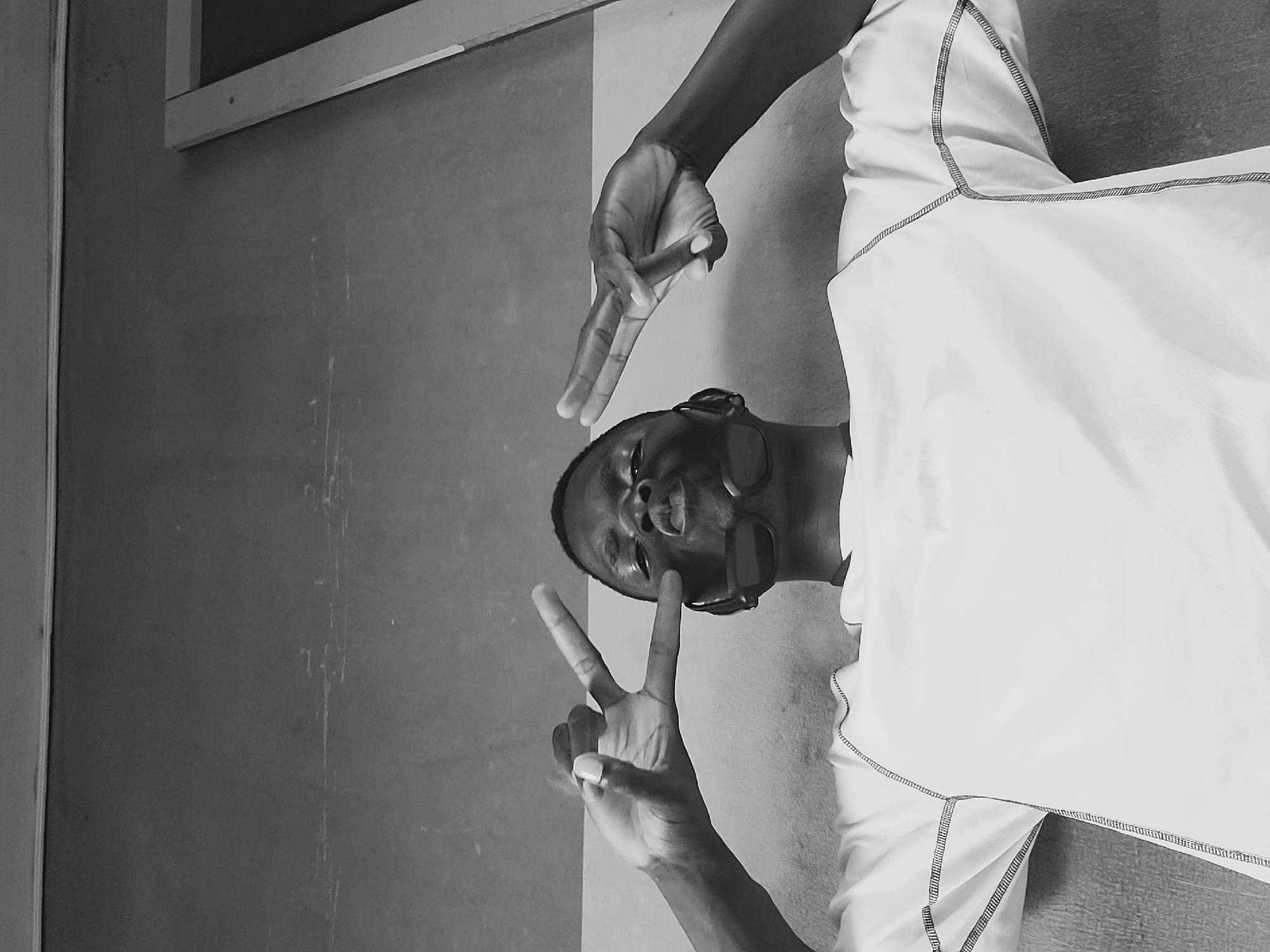 The black and white, low-angle, full shot shows a young Black male wearing a white, long-sleeved shirt. He is in a slightly tilted, dynamic pose, with his head tilted back and his eyes closed, a slight smile on his face. He is wearing sunglasses. His right arm is raised, and his hand is in a peace sign gesture, positioned above his head. His left arm is also raised, with his hand making a peace sign gesture in front of his face. The subject is positioned in the foreground, with a neutral background that appears to be a wall. The lighting suggests it might be daytime, possibly indoors or in a shaded outdoor area. The overall mood of the image is celebratory or playful, with the subject conveying a sense of joy and confidence. The location is identified as Gura Top, Nigeria.