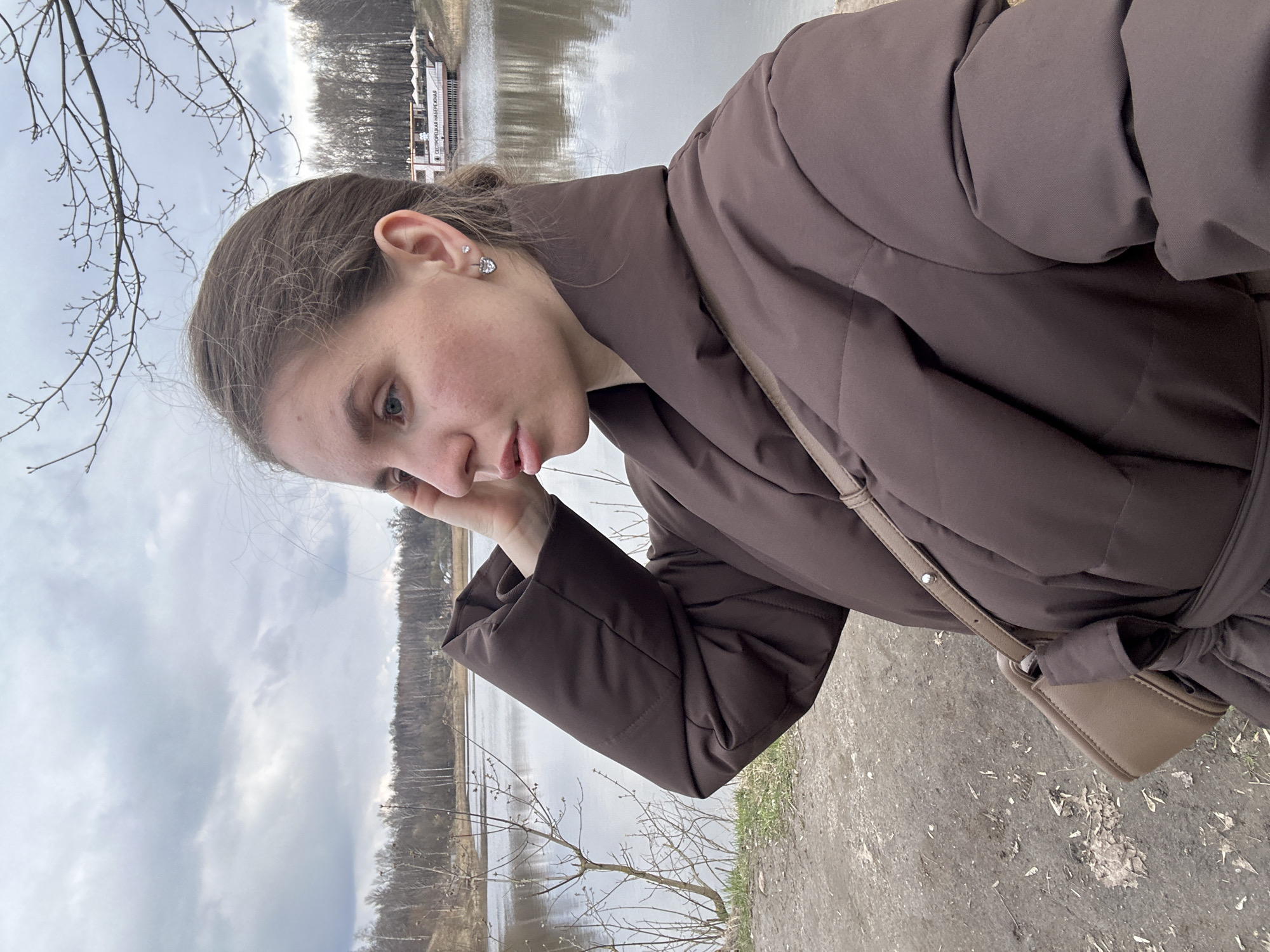 A young woman is depicted in a selfie against an outdoor backdrop. She is wearing a brown, belted puffer jacket and a small beige shoulder bag is visible on her right side. Her hair is pulled back. In the background, a body of water, likely a river or lake, is seen with bare trees along the shoreline, suggesting a colder season, potentially late autumn or early spring. A building is visible on the opposite side of the water. The ground appears to be a dirt path. Given the location context of Klin, Russia, the image is likely taken near the P.I. Tchaikovsky State House Museum located in Klin. The weather conditions appear overcast, indicated by the cloudy sky.