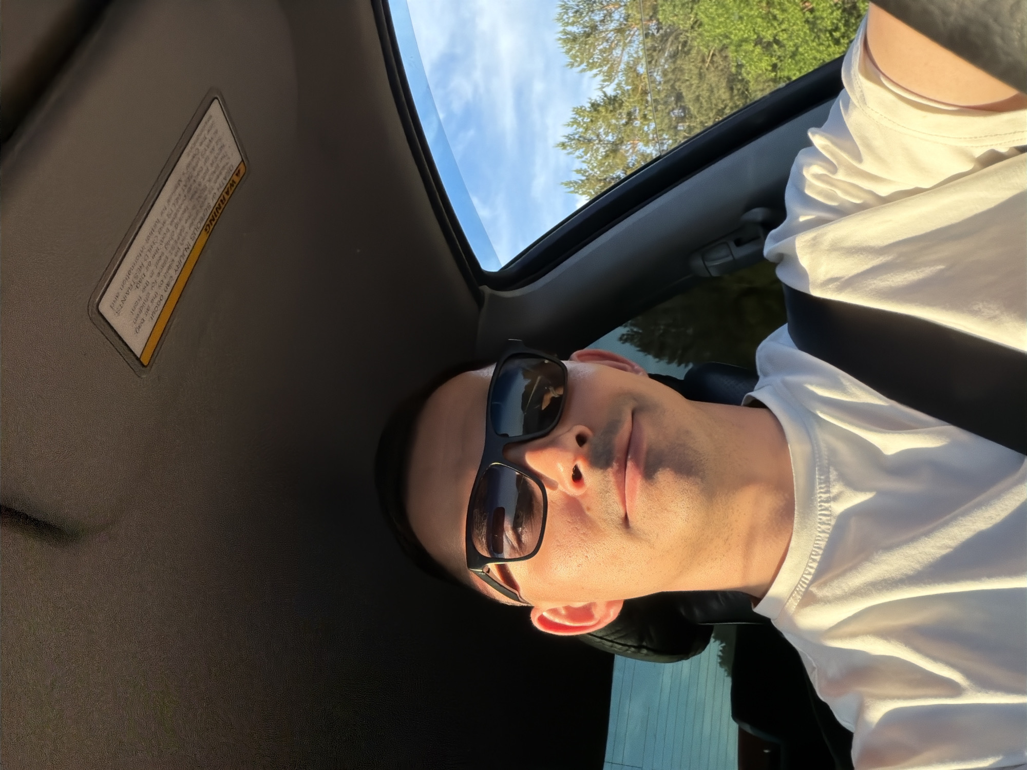 The image captures a person, likely male, from a low angle within a car's interior. He wears black sunglasses and a white t-shirt, looking directly at the camera with a neutral expression. The scene is illuminated by warm, golden sunlight hitting the right side of his face and shirt, suggesting late afternoon or early evening.
Above him, an open sunroof reveals a clear blue sky with sparse clouds and the tops of green trees, indicating an outdoor setting. The car's interior features a dark headliner. A visible warning sticker on the sun visor reads: "A WARNING. DEATH OR SERIOUS INJURY can occur. NEVER put a rearward-facing child seat in the front. The BACK SEAT is the safest place for children. ALWAYS use seat belts and child restraints. See the Car's Manual for further information and safety." The warning emphasizes child safety and proper use of car seats and restraints.
The person appears relaxed while in the vehicle, which is situated outdoors in Yus'ki, Russia, under sunny weather conditions.