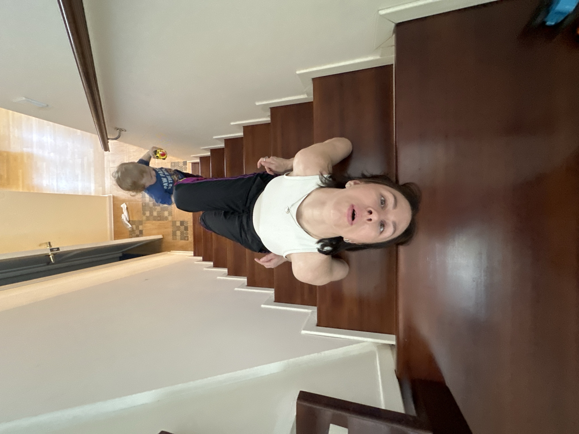A woman in a white tank top and black pants is lying on a set of wooden stairs in what appears to be an indoor setting. In the background, a young child with blonde hair is holding a yellow toy. A partial view of a room with a light-colored floor is visible behind the child. The setting resembles a domestic residence. The location is unknown and there is no reference to Русский Вожой, Russia.