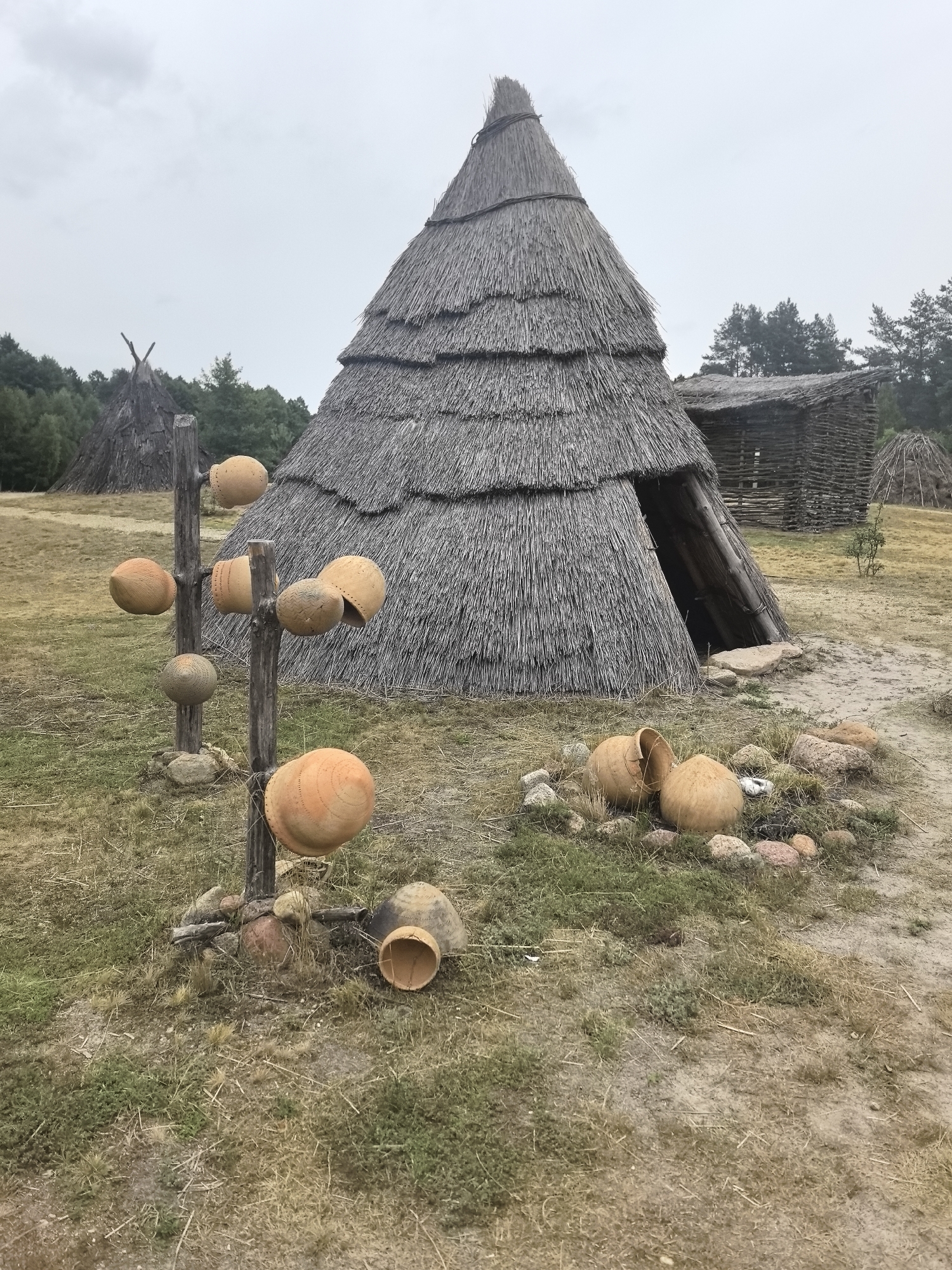 The image depicts an outdoor scene at an archeological reconstruction site in Камянюцкі сельскі Савет, Belarus. In the foreground, a vertical wooden post displays several clay pots of various sizes, some with intricate patterns. These pots are scattered around the base of the post and on the ground. Behind this display, a large conical hut with a thatched roof is prominent. To the right of the conical hut, a rectangular wooden structure with a thatched roof is visible, and further in the background, another conical structure and dense trees line the horizon. The ground is covered with dry grass and patches of soil. The sky is overcast.