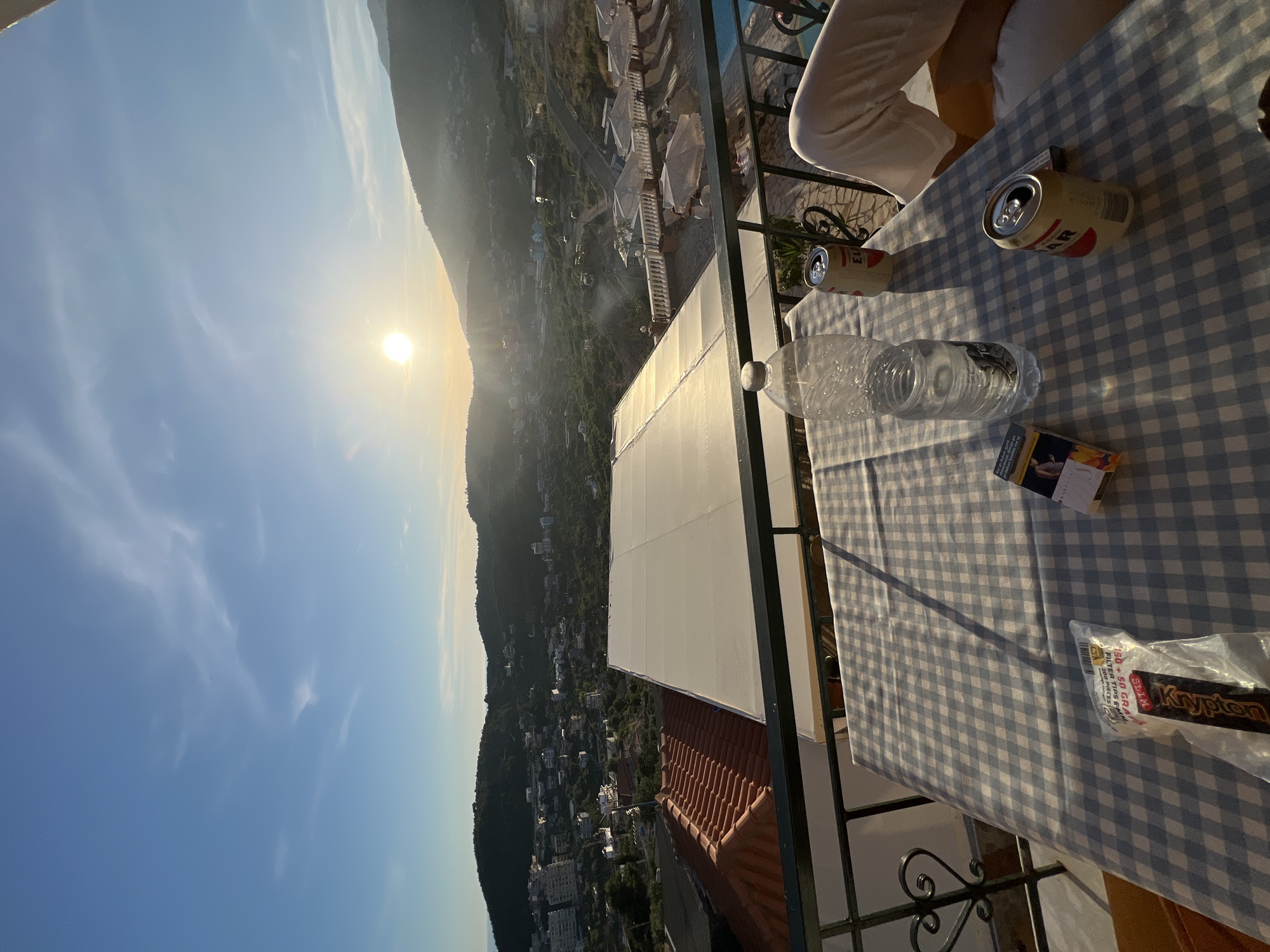 The image depicts a balcony in Himarë, Albania, overlooking a town and hills at sunset. On the table, a checkered tablecloth is set with a plastic water bottle, two opened beer cans, a pack of cigarettes, and a bag of tobacco. A person's legs, clad in white pants, are visible on the right side of the frame. In the background, the sun is setting behind a mountain range, casting a warm glow over the landscape. The town of Himarë, with its buildings and greenery, sprawls across the hills. A swimming pool and lounge chairs are visible further down, suggesting a resort or hotel setting. The sky is a clear blue with a few wispy clouds.