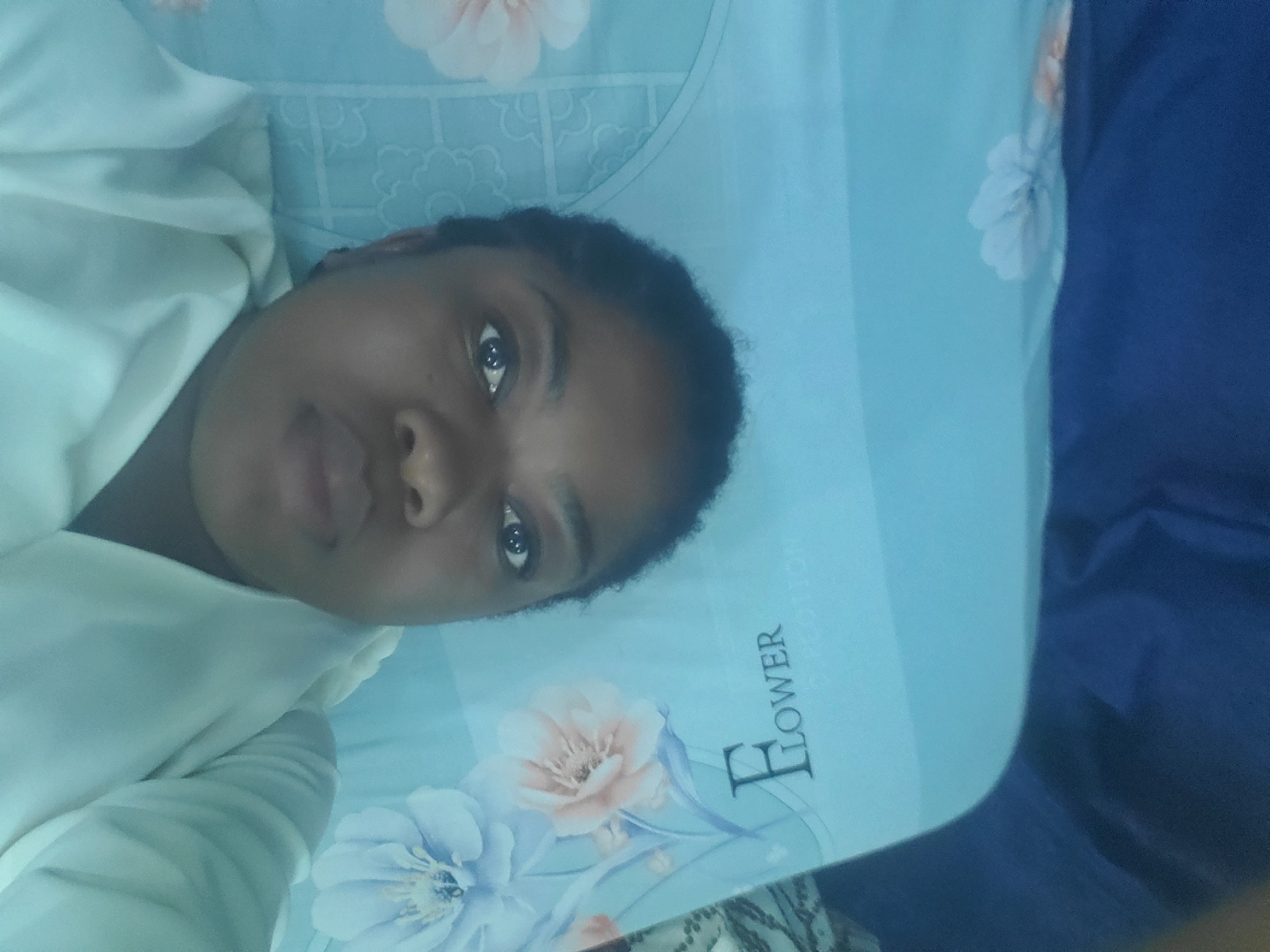 The image features a close-up portrait of a dark-skinned individual, likely a woman, lying down and looking directly into the camera. She has dark, curly hair and a neutral, calm expression. She is wearing a light-colored, possibly white or cream, top with visible folds.
The individual is resting on a light blue bed sheet adorned with large floral patterns in shades of light pink and blue. On the sheet, the word "FLOWER" is clearly visible, written vertically, with the partial letters "TION" appearing below it, suggesting part of the word "COTTON" or a similar material description. To the right of the person, a darker blue fabric, possibly a blanket or another piece of bedding, is visible.
The setting appears to be indoors, likely a bedroom, characterized by soft and even lighting that doesn't indicate a specific time of day. The photograph seems to be a selfie or a casual shot taken from a slightly elevated angle. The scene is situated in Barden Yana, Nigeria.