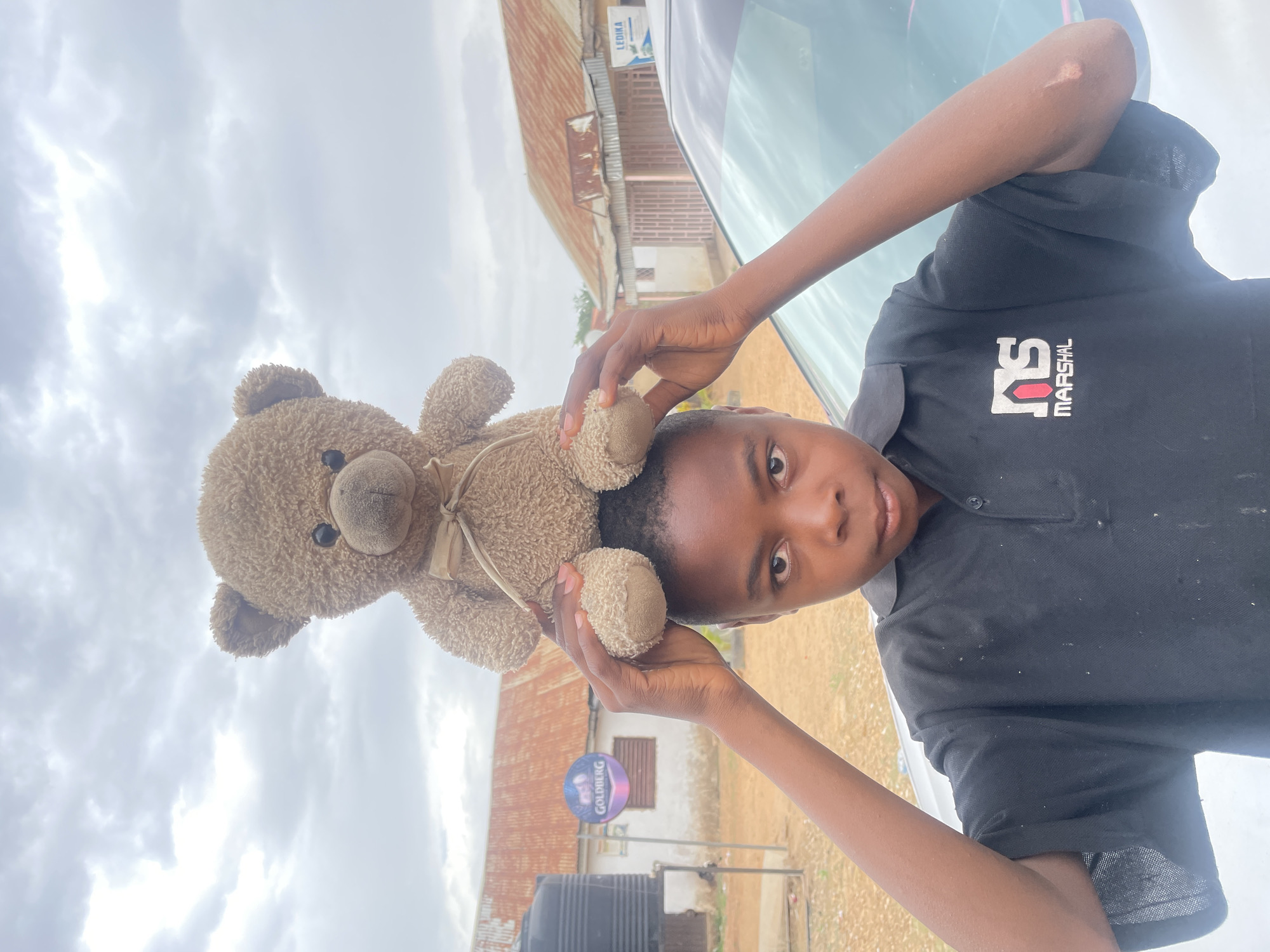 This outdoor image, taken in Barakin Akawo, Nigeria, features a young person holding a teddy bear. The person, with dark skin and short dark hair, is looking directly at the camera with a neutral expression. They are wearing a black polo shirt with a red and white "RS" logo and "MARSHAL" in white text on the left chest. With both hands extended upwards, they hold a brown teddy bear, adorned with a light-colored bow, above their head.
The background indicates a rural or semi-urban setting under a cloudy sky. Visible structures include buildings with reddish-brown corrugated metal roofs and a white wall with a window. A black water tank sits on the ground. In the midground, a prominent round blue and purple sign displays the word "GOLDBERG" in white, accompanied by gold graphics. Another rectangular white sign with blue text reading "LEDIKA" is visible further back, near what appears to be the hood of a silver vehicle. The scene is illuminated by daylight, suggesting an overcast day.