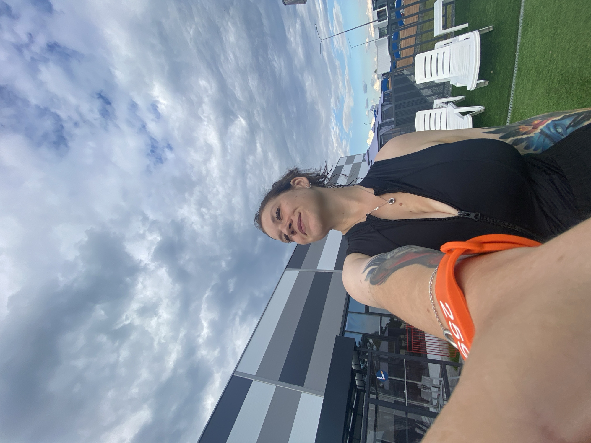 The media file is a selfie taken outdoors in Votkinsk, Russia. The primary subject is a woman with tattoos on her arm, wearing a black zip-up top and an orange bracelet. She is smiling and looking towards the camera. The background shows a modern building with gray and white striped siding, a railing, white lounge chairs, and a patch of artificial turf. The sky is cloudy with patches of blue sky visible, suggesting it might be daytime with variable weather. The overall mood appears relaxed and casual. The orange bracelet on the woman's wrist has white text on it, though it is difficult to read clearly.