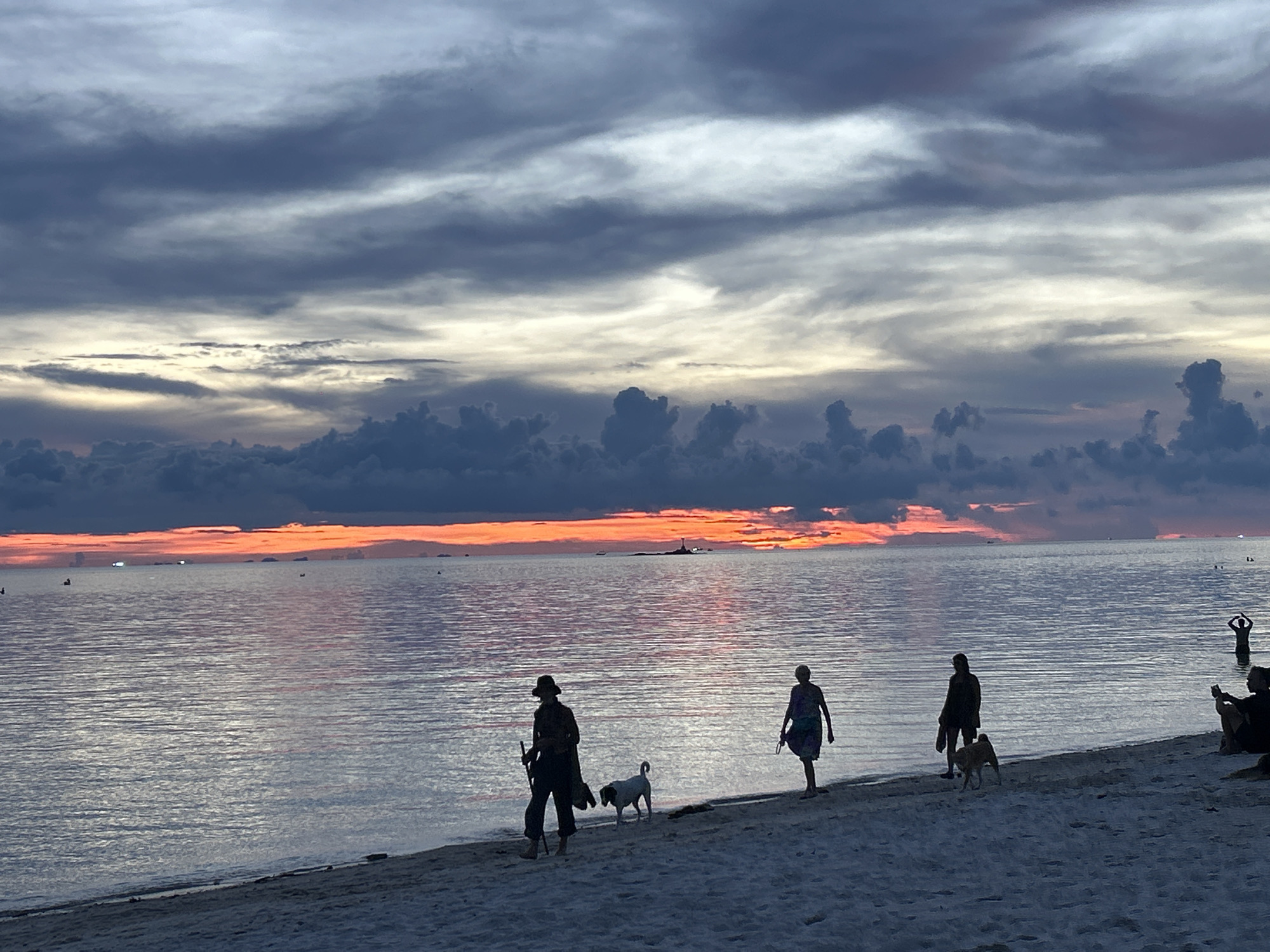 The image captures a serene beach scene at sunset in Baan Sri Thanu, Thailand. The sky is a dramatic mix of dark grey clouds and vibrant orange hues reflecting on the calm ocean. Several silhouetted figures are walking along the shoreline with their dogs. To the right, a person is either in the water or standing near the edge, while a group is seated on the sand. The overall atmosphere is peaceful and evocative of a coastal evening.