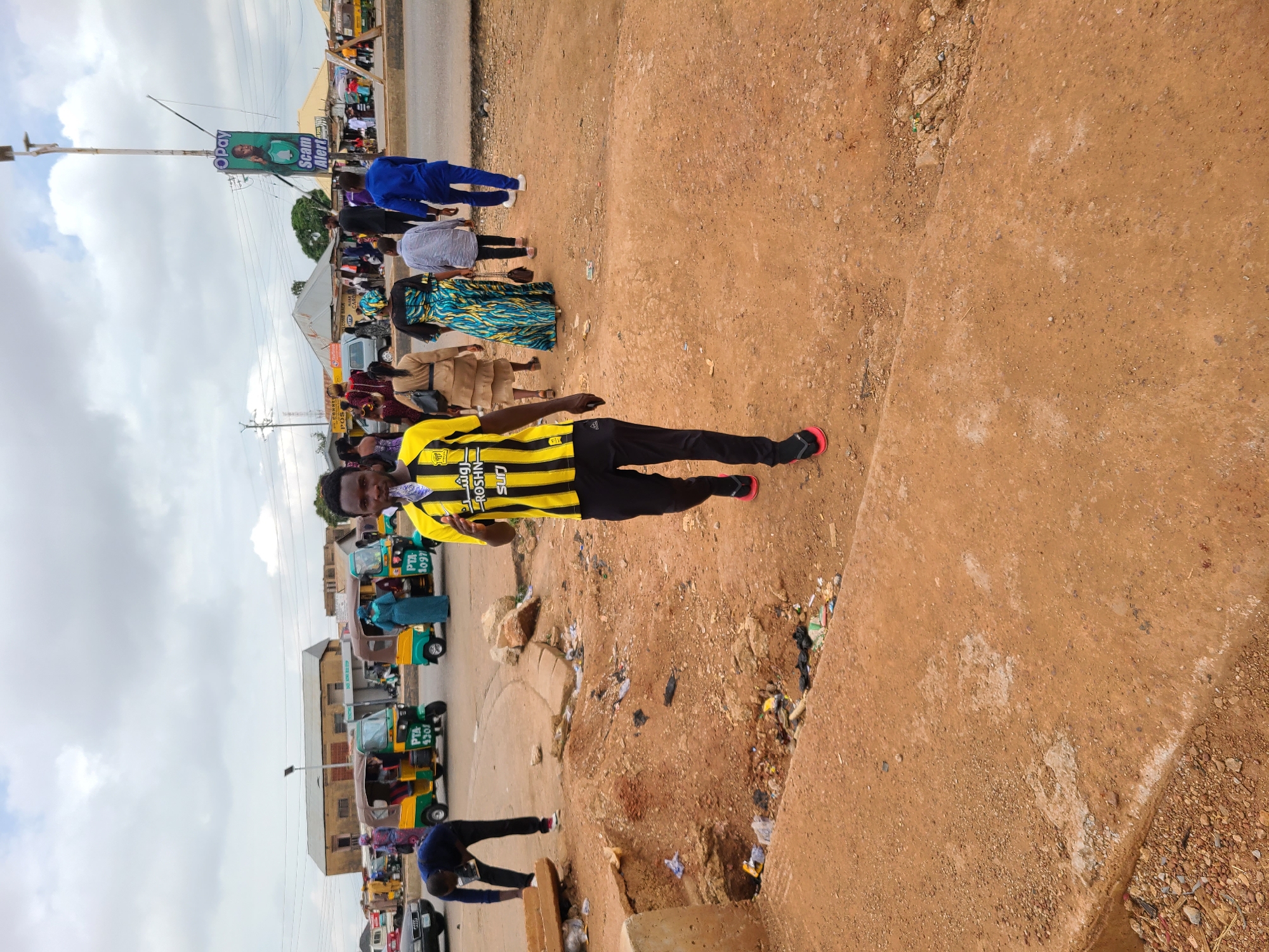 The image shows a street scene with several people and vehicles. A man in a black and yellow striped shirt and black pants is walking, facing the viewer, to the right. Behind him, a line of people are walking in the same direction. There is a row of green and yellow auto rickshaws parked on the left side of the image. Above the people, there is a billboard with an advertisement. The ground is dirt, with some litter and debris. The overall scene appears to be in a city.