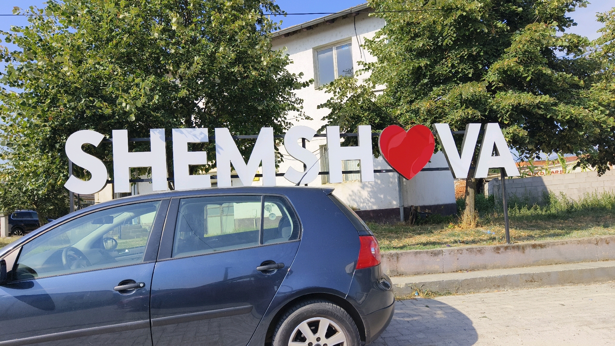 The image shows a sign that reads "SHEMSHEVO" with a red heart symbol in place of the letter "O." The sign is located in front of a white building with a white-tiled roof, surrounded by lush green trees. A dark blue hatchback car is parked in the foreground, partially obscuring the sign. The car has its doors and windows closed. The ground is paved with grey concrete slabs and there is a low wall behind the sign. The sky is clear and blue. The scene appears to be a sunny day.