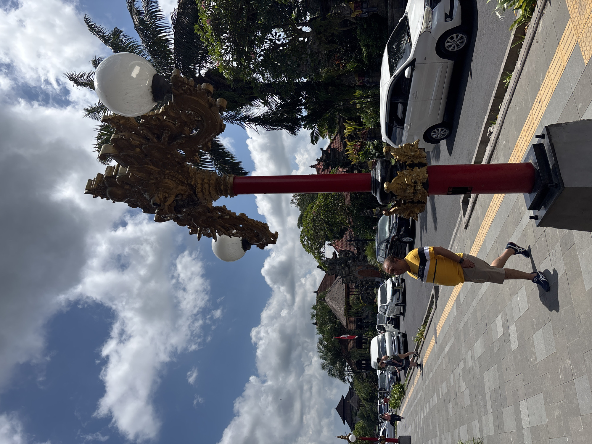 The image depicts a street scene in Ubud District, Indonesia. A middle-aged man is walking along a gray brick sidewalk, wearing a yellow shirt with black and white stripes, khaki shorts, and blue shoes. Cars are parked along the street to his right, including white and black vehicles.
A tall, ornate street lamp with a red pole, gold embellishments, and globe-shaped white lights stands on the sidewalk. Palm trees and other greenery are visible behind the lamp. In the background, traditional Balinese architecture is discernible, including buildings with thatched roofs. The sky is blue with fluffy white clouds.
