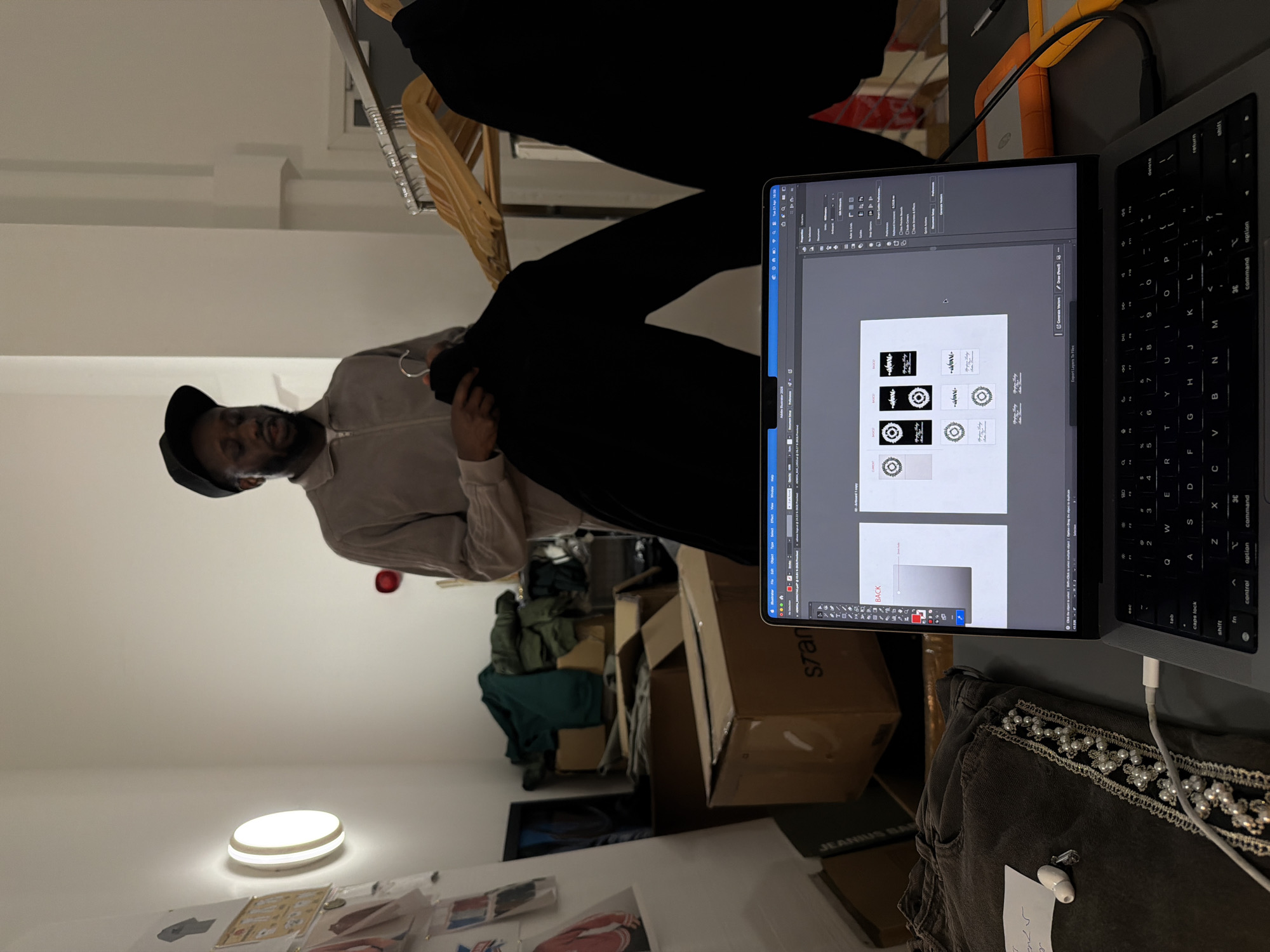 The image, displayed rotated 90 degrees counter-clockwise, depicts an indoor setting, likely a design studio or storage area in London, United Kingdom. A man, positioned sideways on the left side of the frame, wears a light-colored collared shirt and a dark cap. He holds a black garment on a hanger, appearing to inspect it.
To the right of the man, a silver MacBook laptop is open, positioned sideways on a table. Its screen displays Adobe Illustrator 2020, showing various logo designs. These designs are primarily black and white, featuring stylized text and circular motifs, with one design visibly including the text "JEANIUS". The laptop's screen also shows "Tue 23 Apr 16:39", indicating the time. An orange and grey external hard drive is visible behind the laptop. A white charging cable connects to the laptop, near folded dark-colored jeans embellished with pearls and rhinestones, a white earbud, and a piece of paper with handwritten numbers, including "570".
The background features stacked cardboard boxes, also positioned sideways. Some boxes are labeled "sran" and "JEANIUS", suggesting storage or packaging of apparel-related items. Other clothes are visible within some boxes. A circular, illuminated light fixture is mounted on a white wall in the mid-ground, providing artificial light to the room. The overall scene suggests a creative or business environment focused on fashion or branding activities.
Visible text includes: "JEANIUS" and "sran" on boxes; "Adobe Illustrator 2020", "Export Layers To Files", "Generate Vectors", "BACK", and UI elements on the laptop screen; and standard keyboard labels such as "shift", "control", "option", "command", "return", and "delete".